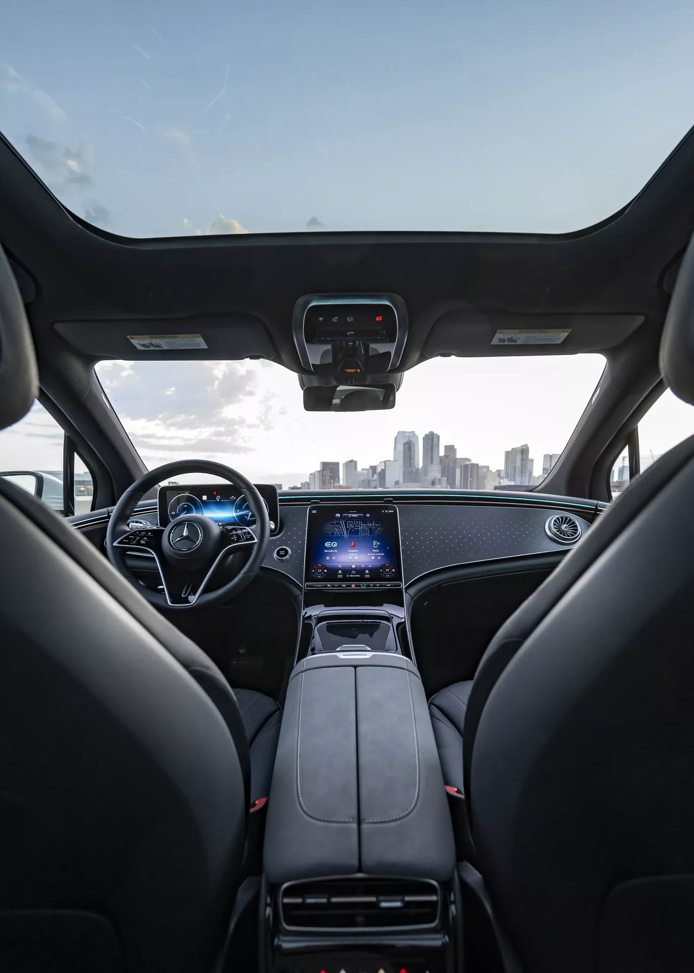 Notranji pogled na Mercedes EQS SUV 2022 iz perspektive zadnjih sedežev, prikazuje sprednje sedeže, armaturno ploščo, volan in vetrobransko steklo. Prednja sedeža sta črn usnjena z vzglavniki, armaturna plošča ima eleganten dizajn s kovinskimi poudarki. Osrednja konzola vsebuje prestavno ročico, držala za pijačo in velik vertikalno orientiran zaslon na dotik za informacijsko-razvedrilni sistem, GPS in nastavitve vozila. Volan vključuje več kontrolnih gumbov, za njim pa je viden digitalen instrumentni prikazovalnik. Slika tudi zajema mestno obzorje skozi vetrobransko steklo, nakazuje vrhunsko, sodobno notranjost z naprednimi tehnologijami.