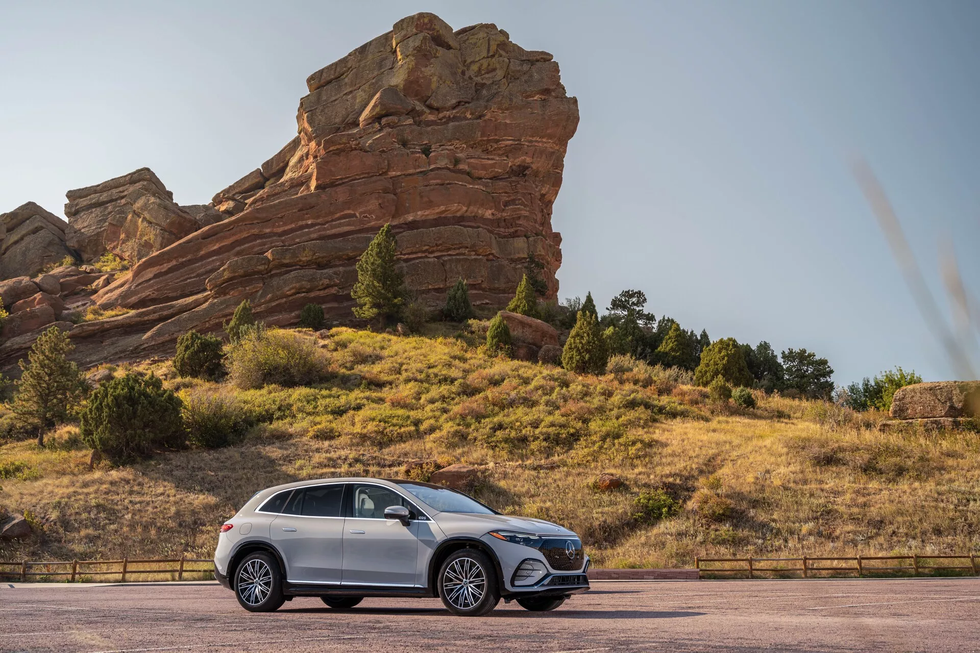 Stranski profilni pogled na Mercedes EQS SUV 2022, parkiran na asfaltirani cesti z velikimi skalnimi formacijami v ozadju, poudarjajoč njegovo večjo oddaljenost od tal, aluminijasta platišča, elegantne oblikovne linije, zatemnjena okna in kovinsko barvano karoserijo.