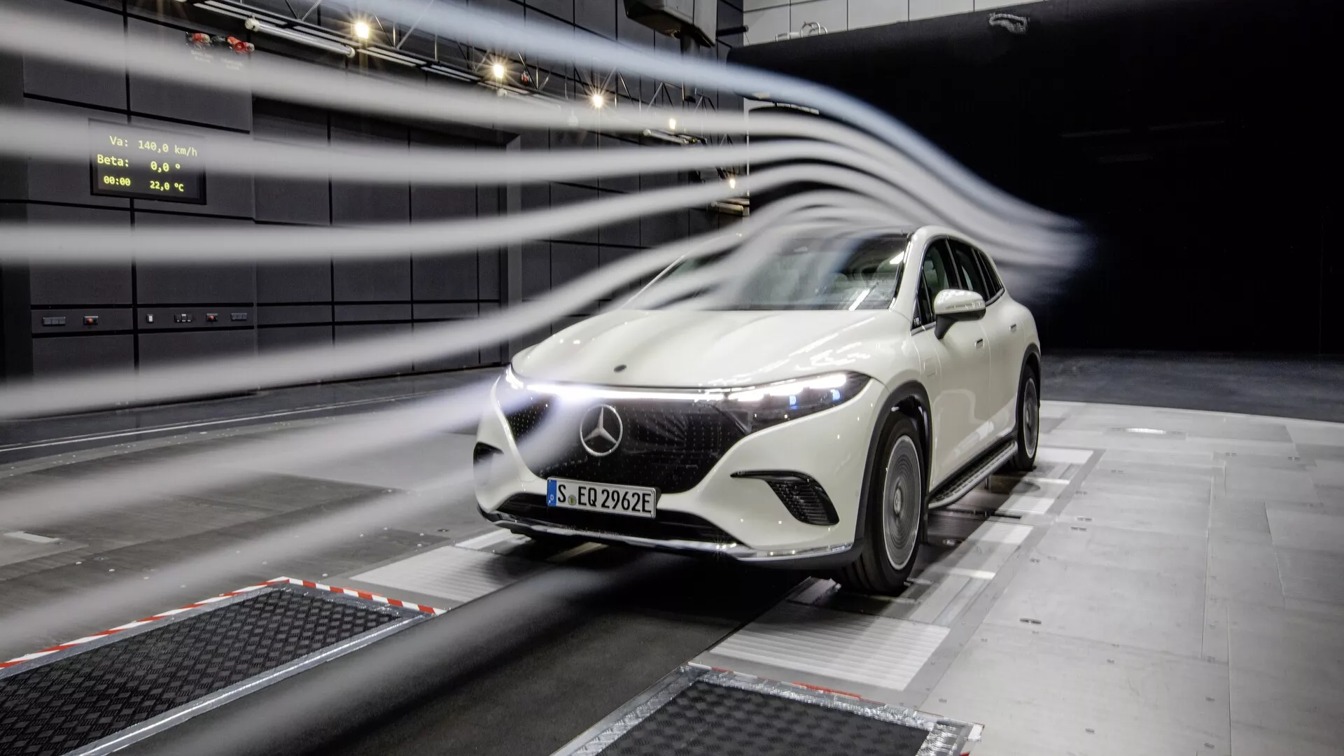 Slika belega Mercedes EQS-SUV 2022 med testiranjem v profesionalnem objektu. Avto je, z rahlim kotom, postavljen v center okvirja in prikazuje svojo sprednjo mrežo, LED žaromete, pokrov motorja, vetrobransko steklo in del strani voznika. Črte zamegljenega gibanja nakazujejo aerodinamične teste. Futuristična oblika, velik emblem na mreži in notranje testno okolje.