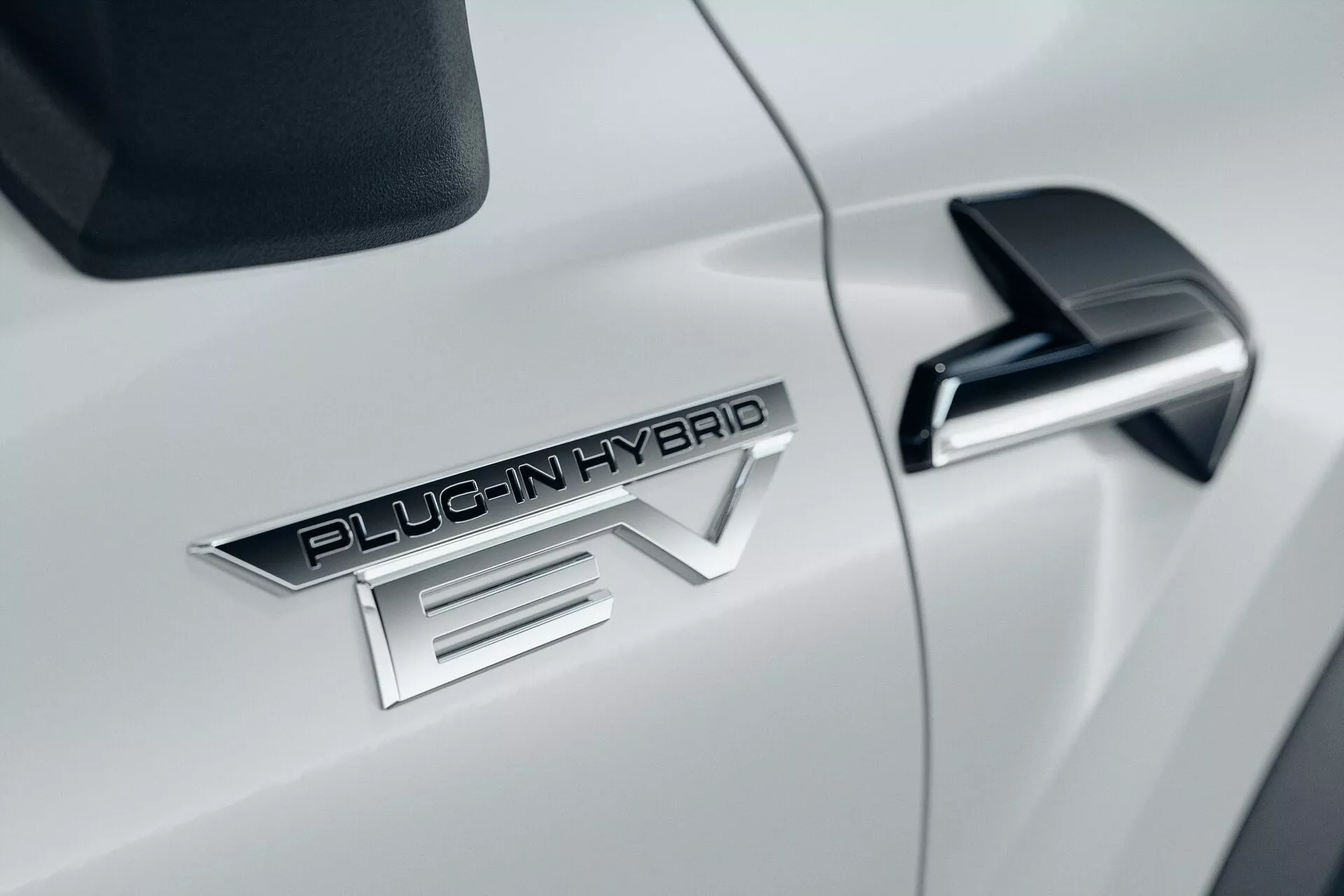 Bližnji posnetek strani belega avtomobila Mitsubishi ASX 2023 druge generacije, z vidnim znakom 'PLUG-IN HYBRID EV', elegantnim črnim stranskim ogledalom z vgrajenim smernikom in sijočo belo aerodinamično karoserijo.