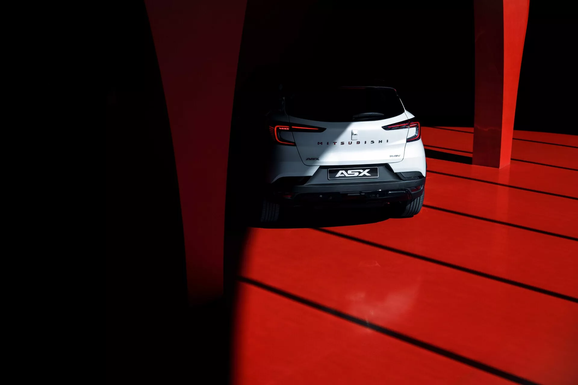 Posnetek iz nizkega kota zadnjega dela kompaktnega SUV Mitsubishi ASX 2023 z izoblikovanimi zadnjimi lučmi, kromiranim znakom in zadnjim vetrobranskim steklom, zajet ob močnem dnevnem kontrastu na živordeči površini.