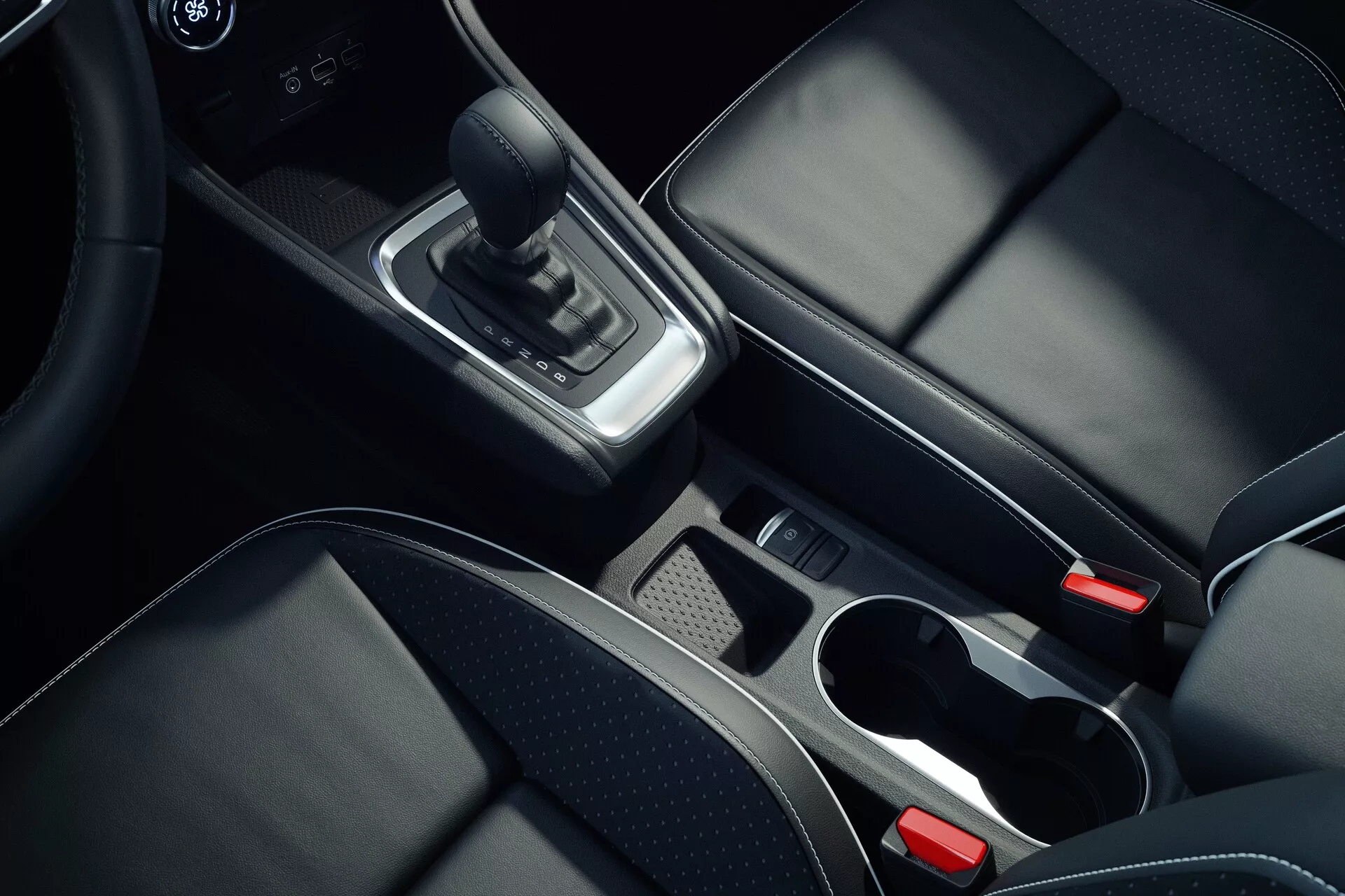 Notranja slika sredinske konzole avtomobila Mitsubishi ASX 2023 druge generacije, posneta z zgornje perspektive pod kotom približno 45 stopinj, osredotočeno na podrobnosti, kot so usnjen menjalnik, kromirani okraski okoli območja prestavne ročice, predal za shranjevanje z odstranljivo pladnje, dva nosilca za kozarce s kromiranimi robovi, črni usnjeni sedeži s vidnimi šivi, gumb za elektronsko parkirno zavoro in delno vidna sredinska naslonjala ter sistem za informacijsko-zabavni ali klimatsko napravo, kar prikazuje visokokakovostne materiale in sodoben dizajn.