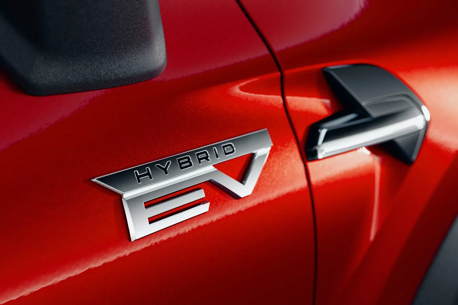 Podroben posnetek rdečega vozila Mitsubishi ASX 2023 (2. generacija), ki poudarja 'HYBRID EV' značko, kar označuje tehnologijo hibridnega električnega vozila. Značka 'HYBRID EV' je v središču, poudarjajoč kovinski zaključek in natančne črke. 'EV' ima značilno puščico, ki prečka 'V'. Stransko ogledalo je delno vidno v zgornjem desnem kotu, kar nakazuje na dodatne tehnološke funkcije. Sijajna končna obdelava laka in tesen šiv med sovoznikovimi vrati in sprednjim blatnikom nakazujeta odlično izdelavo.