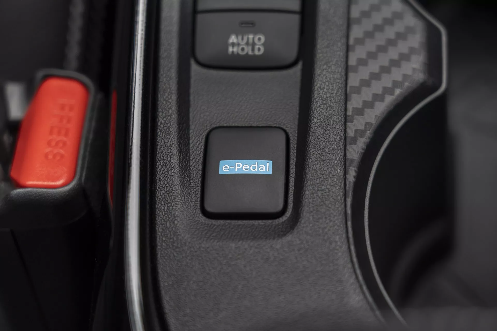Podrobni posnetek notranjosti Nissan Juke (F16) 2019 s poudarkom na stikalu sistema e-Pedal za nadzor električnega ali hibridnega pogona, z vidnim gumbom AUTO HOLD in rdečo zaponko varnostnega pasu, kar nakazuje napredno avtomobilistično tehnologijo in premium notranje funkcije