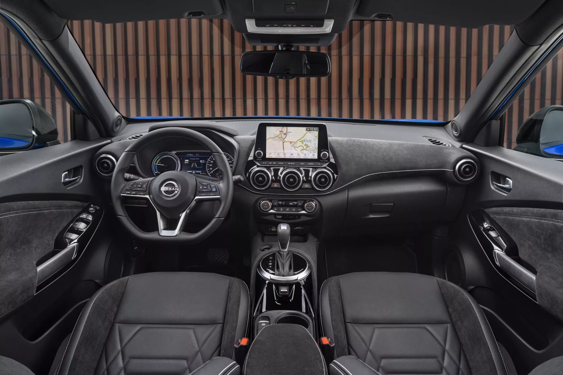 Notranji pogled Nissan Juke (F16) 2019 iz zadnjega sedeža, ki prikazuje volan na levi strani z nadzornimi gumbi, osrednji zaslon na dotik z navigacijo, ročni menjalnik, sedeže z diamantnim vzorcem šivanja in nadzor sončne strehe
