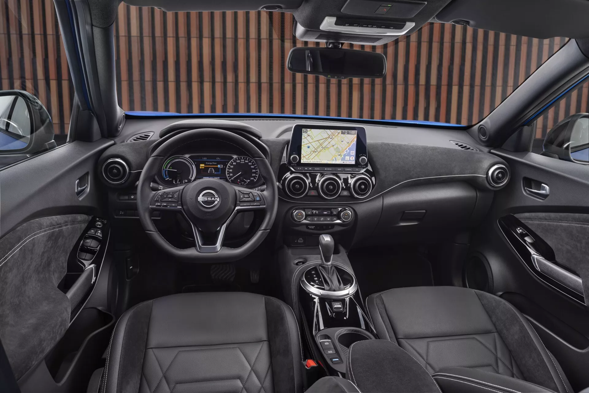 Notranjost Nissan Juke F16 iz leta 2019 iz perspektive voznika, z modernim volanom s tremi kraki z nadzornimi gumbi, kombinirano digitalno in analognim instrumentnim zaslonom, centralnim zaslonom na dotik z navigacijo, dvopodročno klimatsko kontrolo, avtomatskim menjalnikom, usnjenimi sedeži v obliki vedra in elektronsko parkirno zavoro.