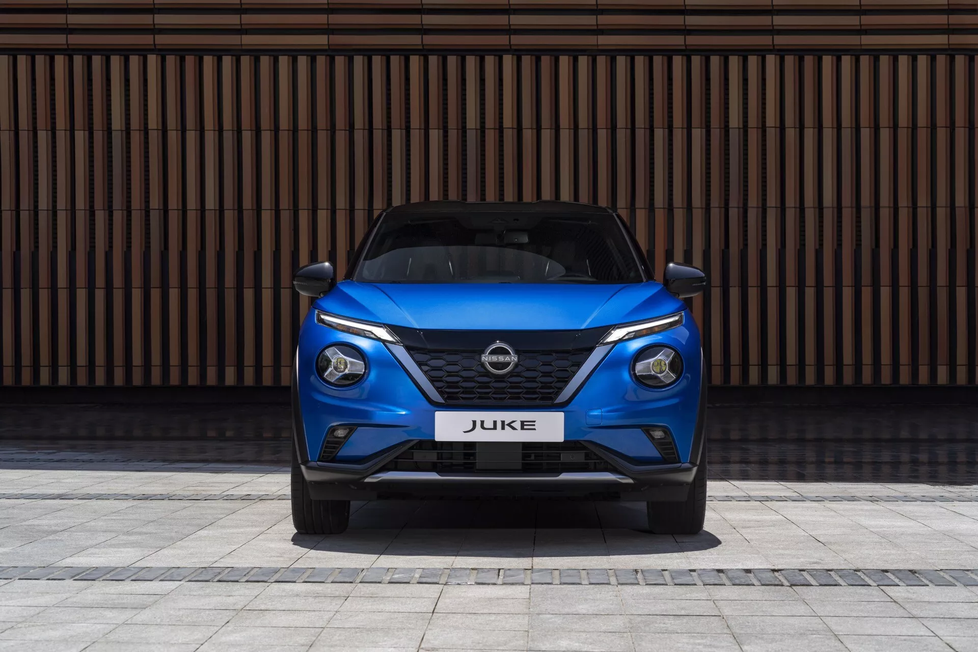 Sprednji pogled na modri Nissan Juke F16 iz leta 2019, ki prikazuje elegantne LED žaromete, V-motion masko in športni odbijač na ozadju z zidom s horizontalnimi linijami
