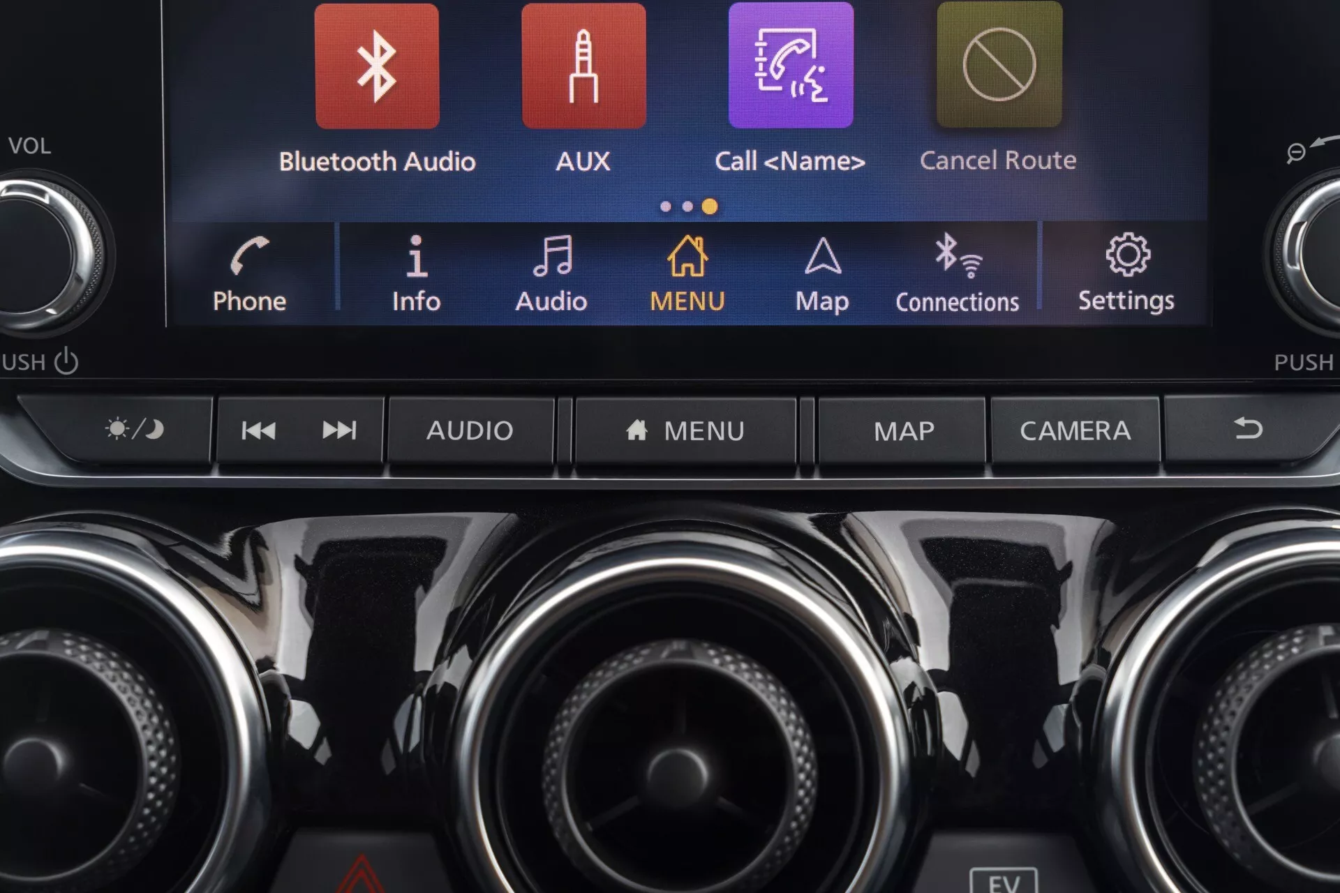 Podroben pogled na osrednjo konzolo Nissan Juke (F16) 2019 z zaslonom na dotik za informacijsko-razvedrilni sistem, ki prikazuje barvne ikone, kot so Bluetooth Audio, AUX in Zemljevidi, skupaj z gumbi za nadzor klime in dodatnimi funkcionalnimi gumbi, poudarjajoč sodobno tehnologijo vozila in premijsko notranjo opremo