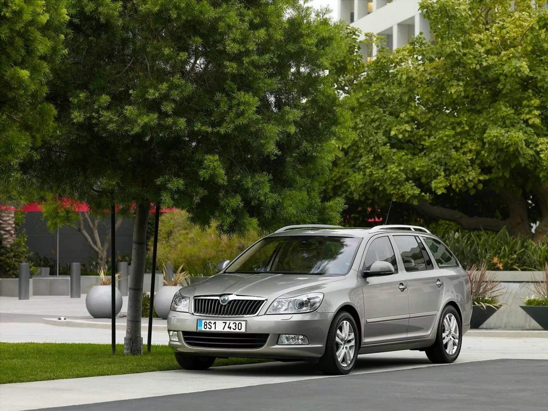 Opel Astra karavan letnik 2011 parkiran v urbanem okolju z zelenjem in modernimi stavbami v ozadju, prikazuje sprednjo rešetko avtomobila, žaromete in eleganten bočni profil, vključno z litoželeznimi kolesi in strešnimi nosilci