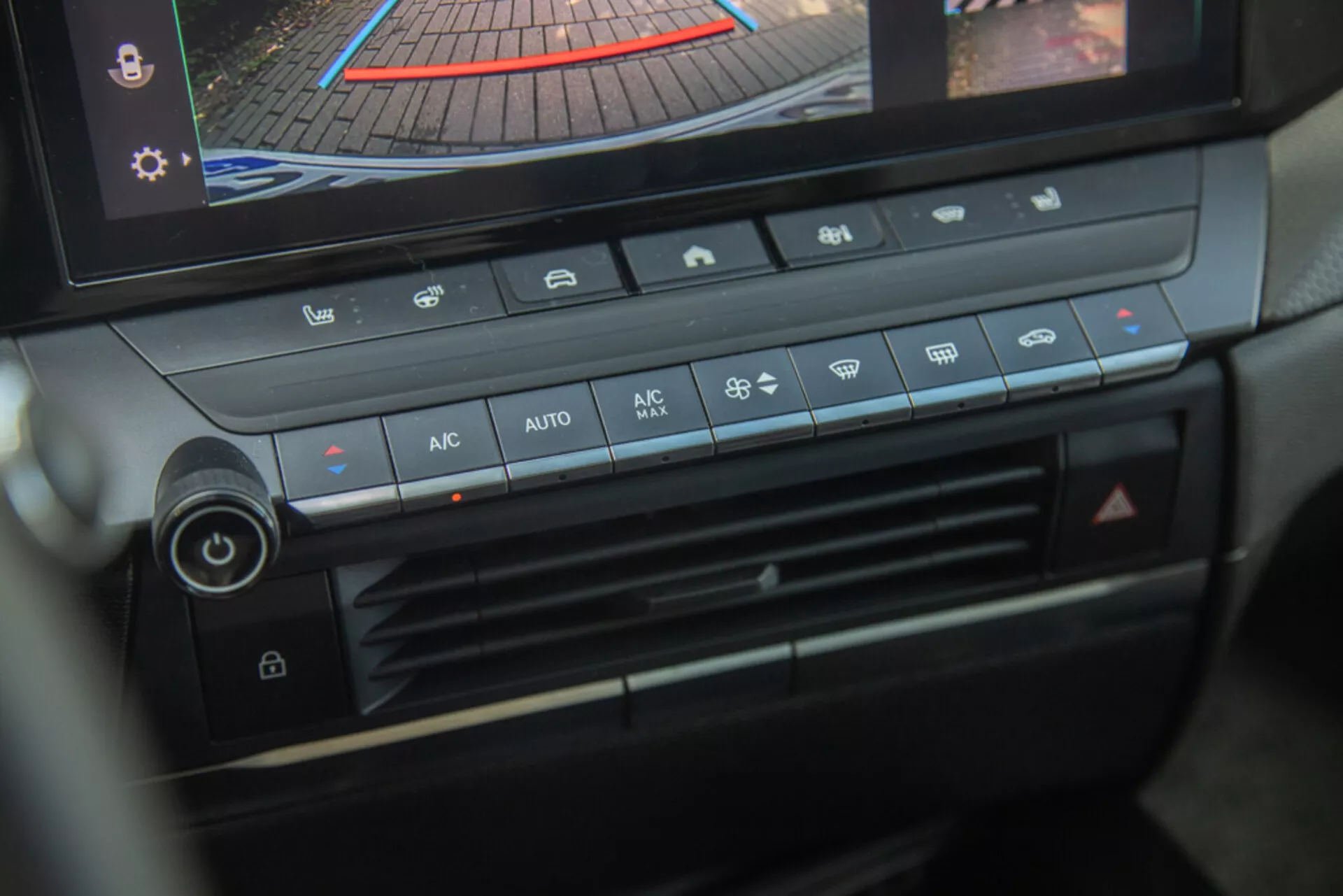Sredinska konzola in plošča za upravljanje klime Opel Astra 2022, poudarjajoč gumb za nastavitev temperature, hitrost ventilatorja, distribucijo zraka in gumbe za avtomatsko klimatsko napravo z digitalnim zaslonom, ki prikazuje pogled kamere za vzvratno vožnjo