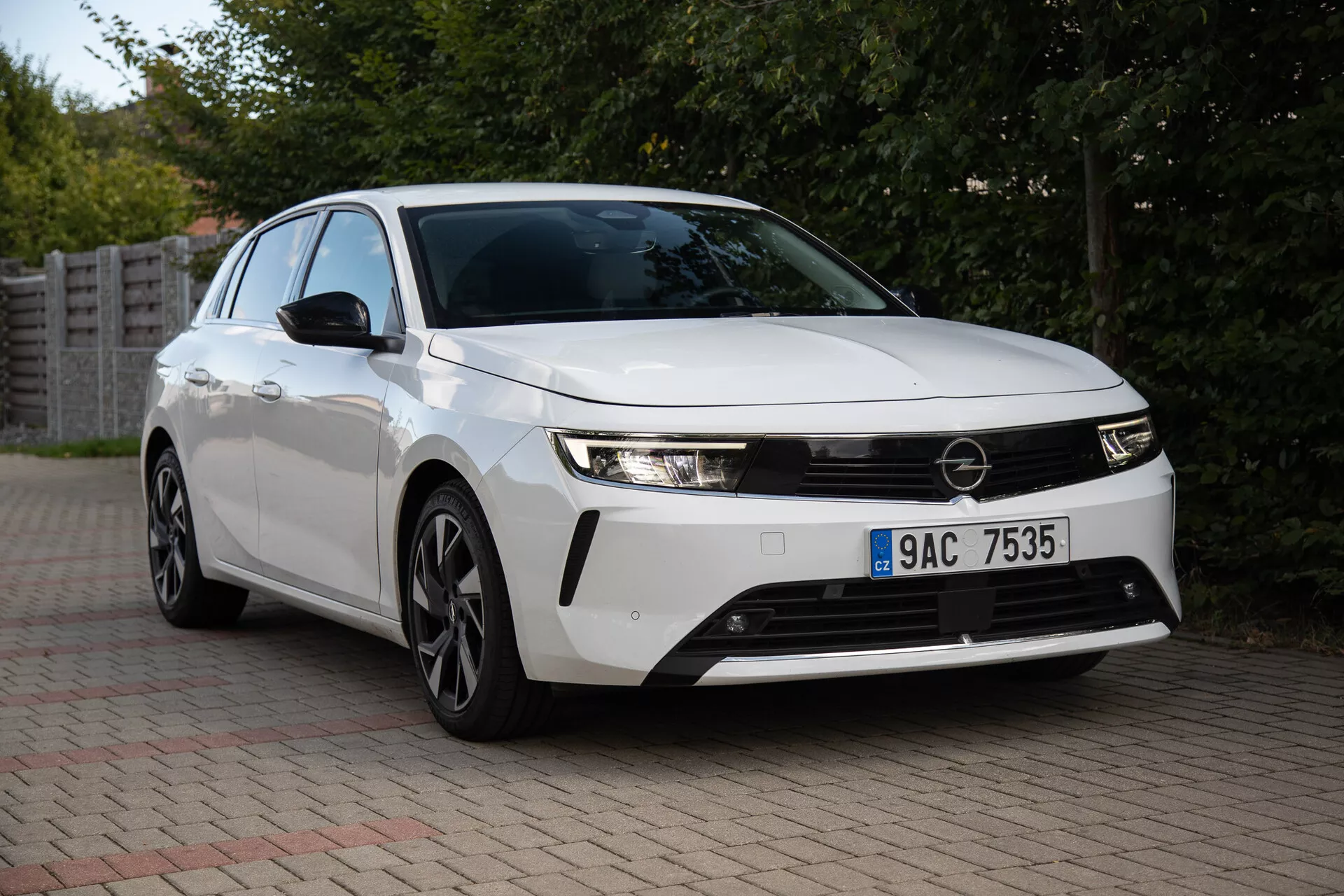 Opel Astra 2022 zajeta v predmestnem okolju, ki prikazuje njene elegantne žaromete, izstopajočo sprednjo masko, aerodinamično obliko vključujoč večkraka lita kolesa in inovativno tehnologijo iz pogleda spredaj pod kotom treh četrtin