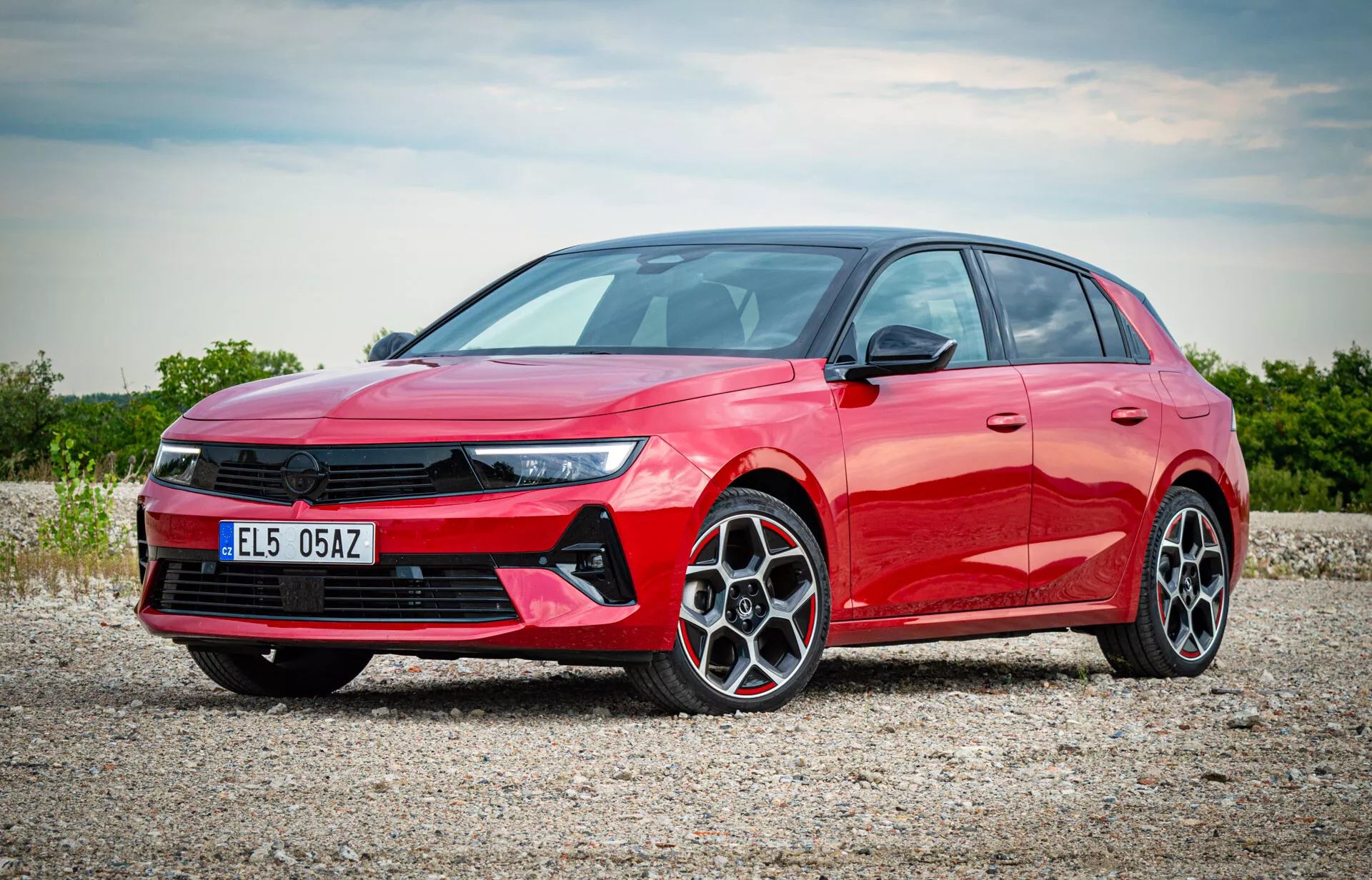 Rdeči hatchback Opel Astra 2022, fotografiran iz pogleda spredaj treh četrtin, ki prikazuje LED žaromete, izstopajočo sprednjo masko, večkraka lita kolesa in elegantni stranski profil na gramozni površini z difuzno sončno svetlobo, ki osvetljuje sodobne značilnosti avtomobila