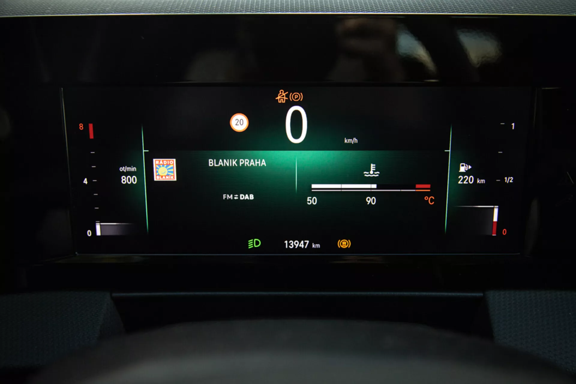Pogled na digitalno armaturno ploščo Opel Astra 2022, ki poudarja instrumentno ploščo z vrtljaji motorja, nivojem baterije, hitrostomerom, merilnikom goriva, zunanjo temperaturo, radijsko postajo 'BLANIK PRAHA' in odčitkom števca prevoženih kilometrov iz perspektive voznikovega sedeža.