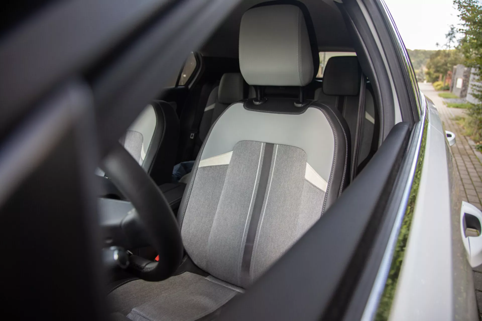Podroben pogled na notranjost voznikove strani Opel Astre 2022 iz zunanje stranskega kota, ki prikazuje dvobarvno voznikovo sedež, integriran naslonjalo za glavo, del volana z kontrolniki, pogled na armaturno ploščo, pripet varnostni pas, notranjost vrat z morebitnimi kontrolniki električnih oken in ohišje stranskega ogledala v barvi karoserije, s poudarkom na ergonomiji, varnosti in funkcionalnosti udobja.