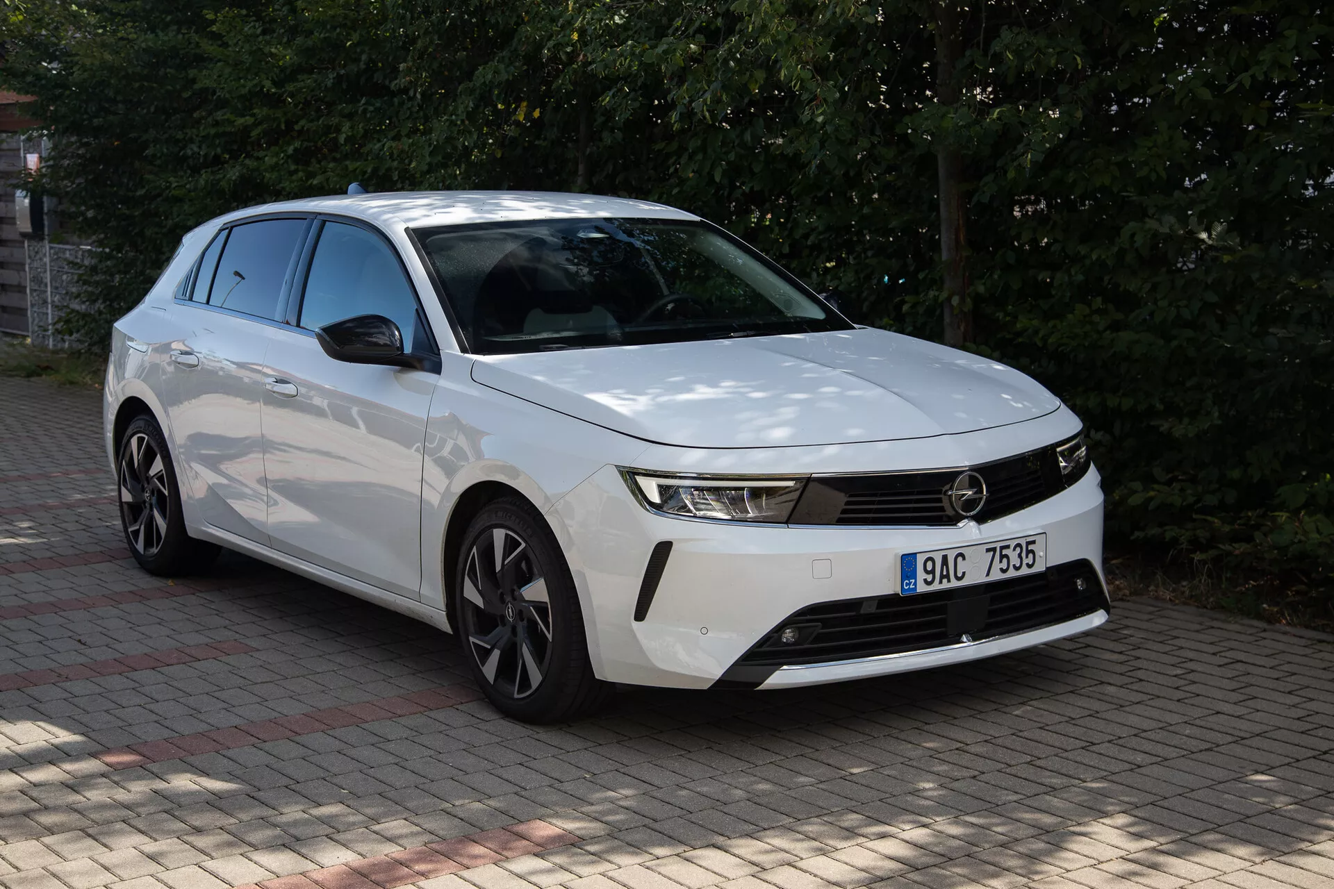 Bela Opel Astra 2022 z izrazitim sprednjim delom, vitkimi žarometi, horizontalno rešetko, aerodinamičnim odbijačem, oblikovanim pokrovom motorja in platišči iz lahke zlitine, posneta iz zornega kota tri četrtine spredaj na prostem podnevi