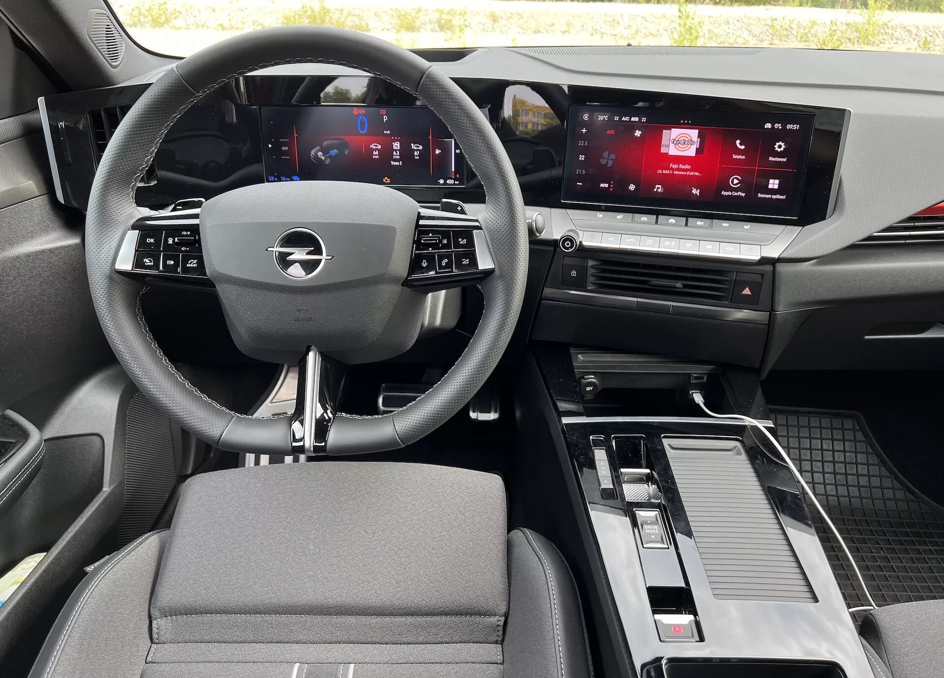Notranjost Opel Astre 2022 iz perspektive zadnjega sedeža, ki prikazuje volan s tremi kraki z integriranimi gumbi, digitalni instrument sklop, zaslon na dotik za informacijsko-zabavni sistem, sredinsko konzolo z ročico menjalnika in prostorom za shranjevanje ter mešanico materialov iz tkanine in videza kovine