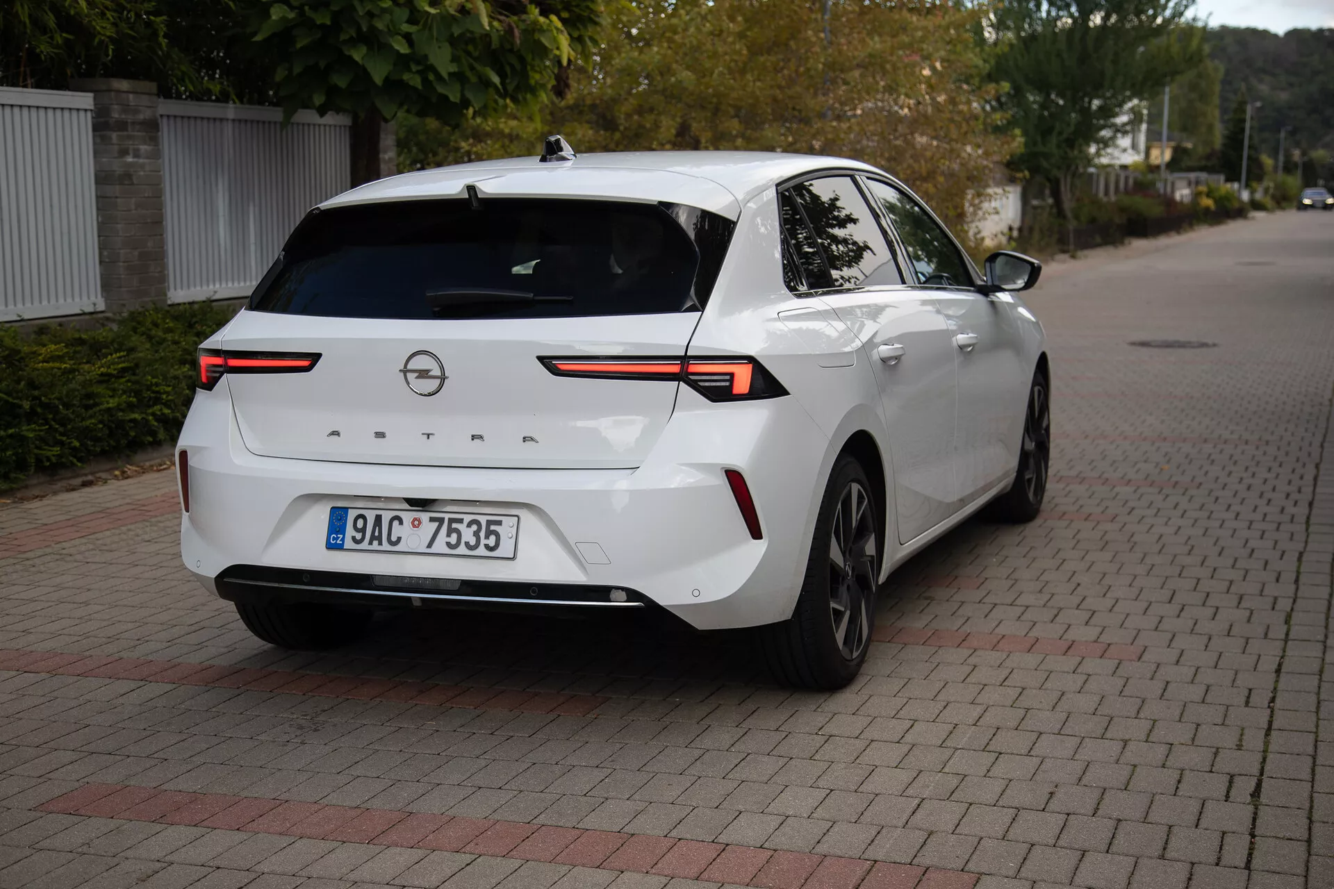 Moderen sedan Opel Astra 2022 v beli barvi, parkiran na površini iz prepletenih opek, prikazuje zadnji tričetrtinski kot z osredotočenjem na visoko tehnološke LED zadnje luči, zadnjo kamero, senzorje za parkiranje, delni stranski profil vključno z desnim zadnjim kolesom in vrati, povezane s horizontalno svetlobno letvijo čez pokrov prtljažnika, opremljene z anteno v obliki morskega psa za izboljšano povezljivost, primerne za ilustracijo zadnjega dizajna vozila in moderne avtomobilske tehnologije