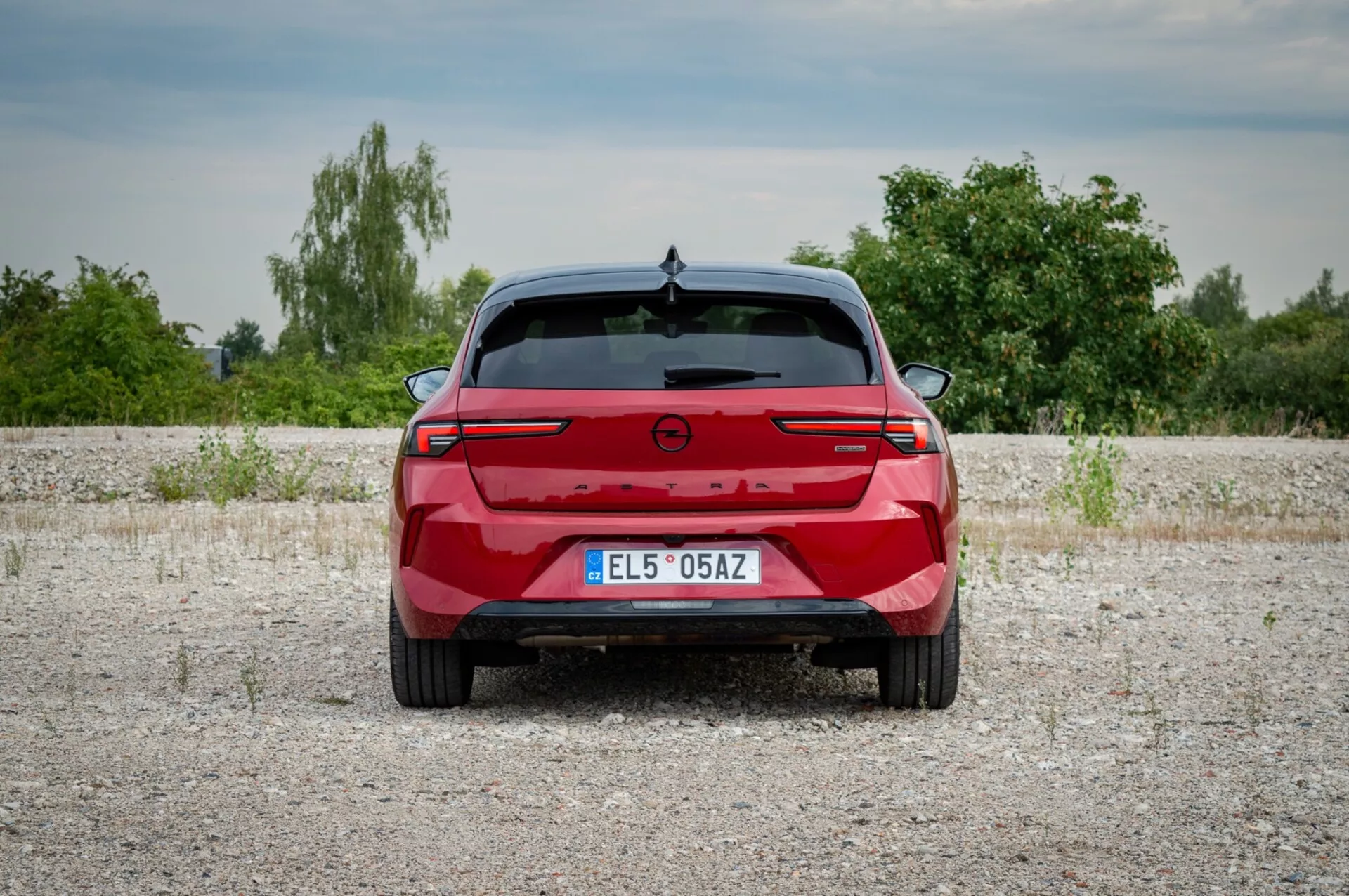 Opel Astra 2022, fotografirana od zadaj na gramozni površini, prikazuje zadnje luči s povezovalno svetlobno letvijo, senzorji za parkiranje in odsotnostjo izpušnih portov, kar nakazuje potencialni električni ali hibridni model, na ozadju vegetacije in rahlo oblačnega neba.