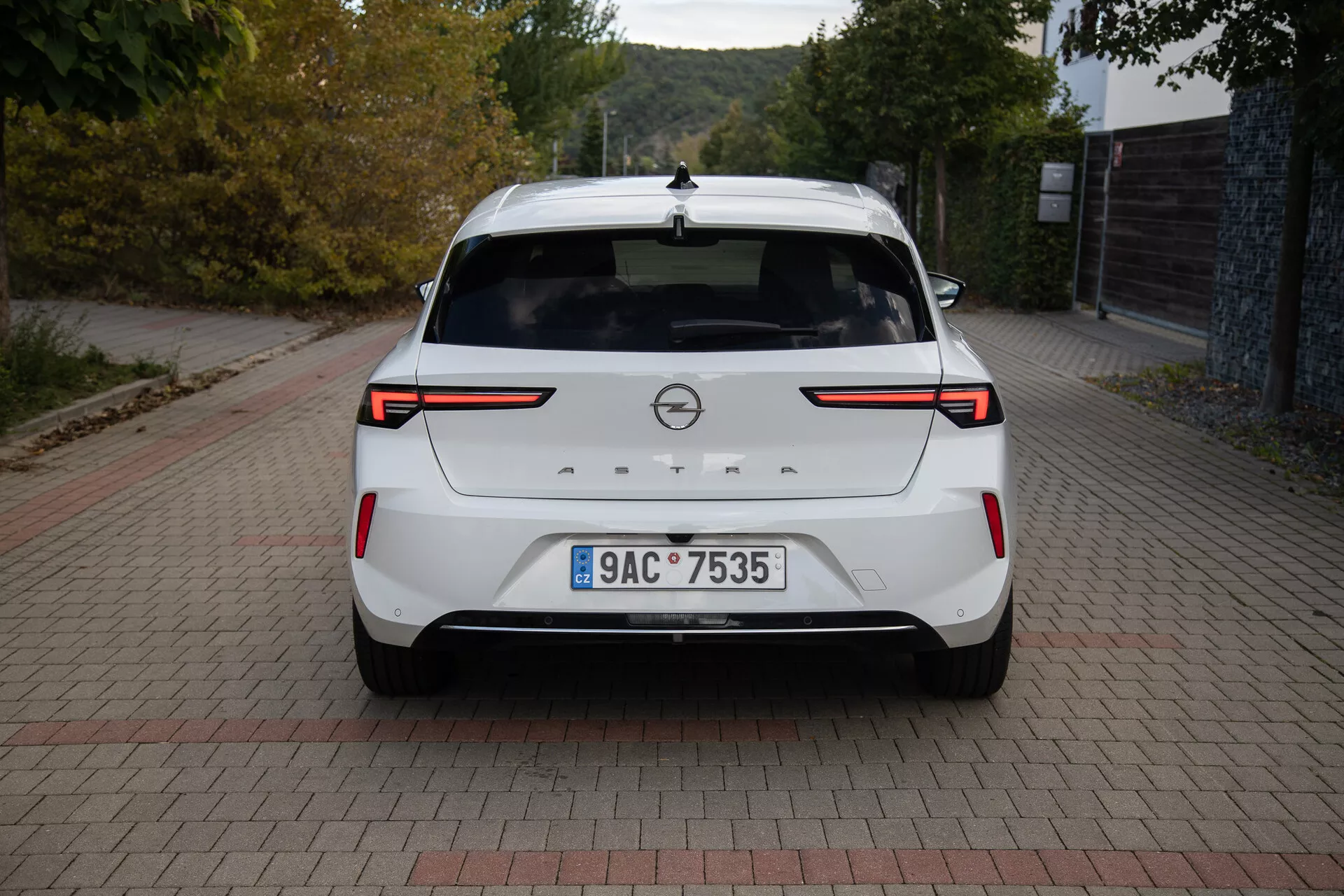 Zadnji pogled na belo Opel Astra 2022, ki prikazuje LED zadnje luči, senzorje / kamero za parkiranje zadaj, znamko proizvajalca, registrsko tablico, zadnji spojler, visoko nameščeno zavorno luč, anteno v obliki plavuti morskega psa in čisto lakirano površino, parkirano na površini opečnatega vzorca podnevi.