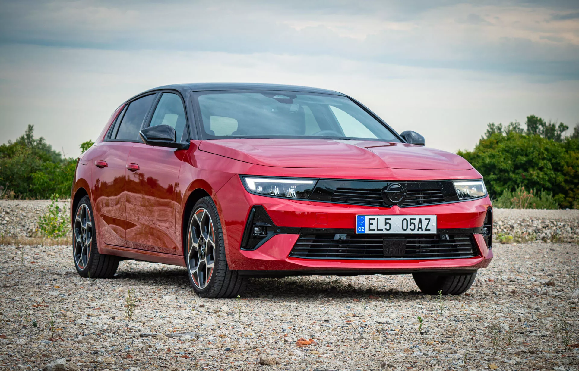 Opel Astra 2022 v živahni rdeči barvi, prikazuje sodobno zasnovo z kotnimi LED žarometi, izstopajočo sprednjo masko, športnim sprednjim odbijačem in večkrakimi zlitimi kolesi, predstavljen v naravnem zunanjem okolju iz pogleda spredaj treh četrtin, ki poudarja aerodinamični profil in premium lastnosti avtomobila