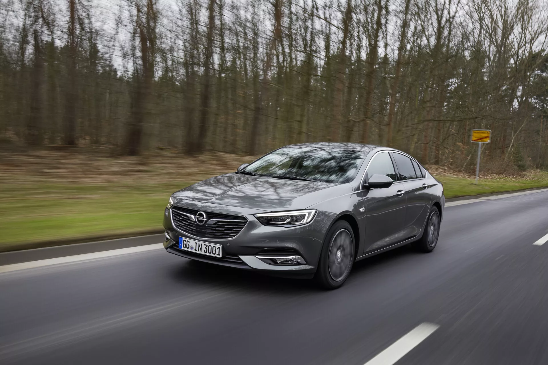 Sodobna limuzina Opel Insignia 2013 v gibanju po ovinkasti cesti skozi gozd, prikazuje stranski profil z LED žarometi, veliko rešetko, lita kolesa, stranska ogledala z indikatorji in zatemnjenimi stekli
