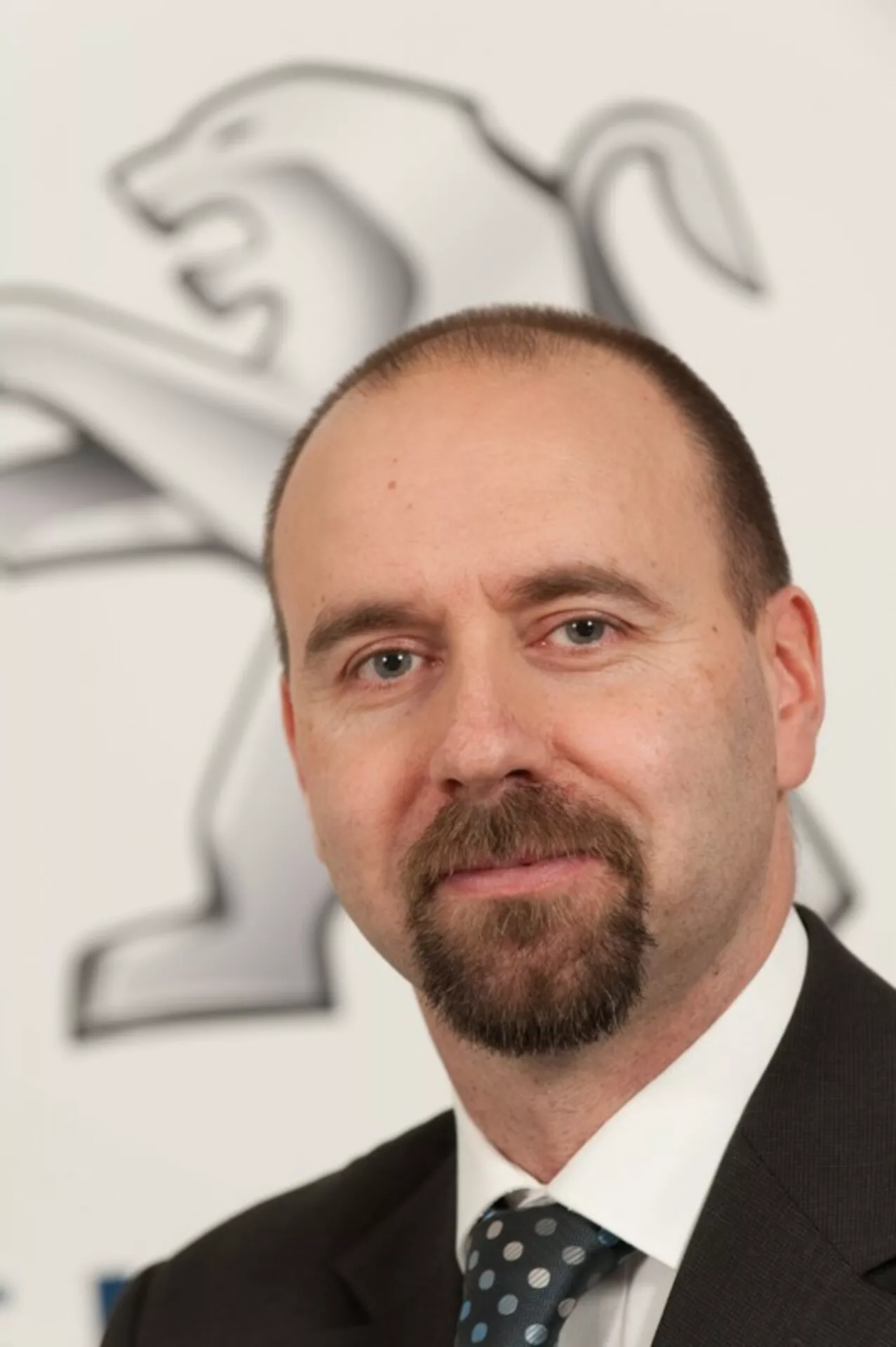 Profesionalni portret moškega v obleki in kravati z logotipom Peugeot (lev) v ozadju, avto ni prikazano