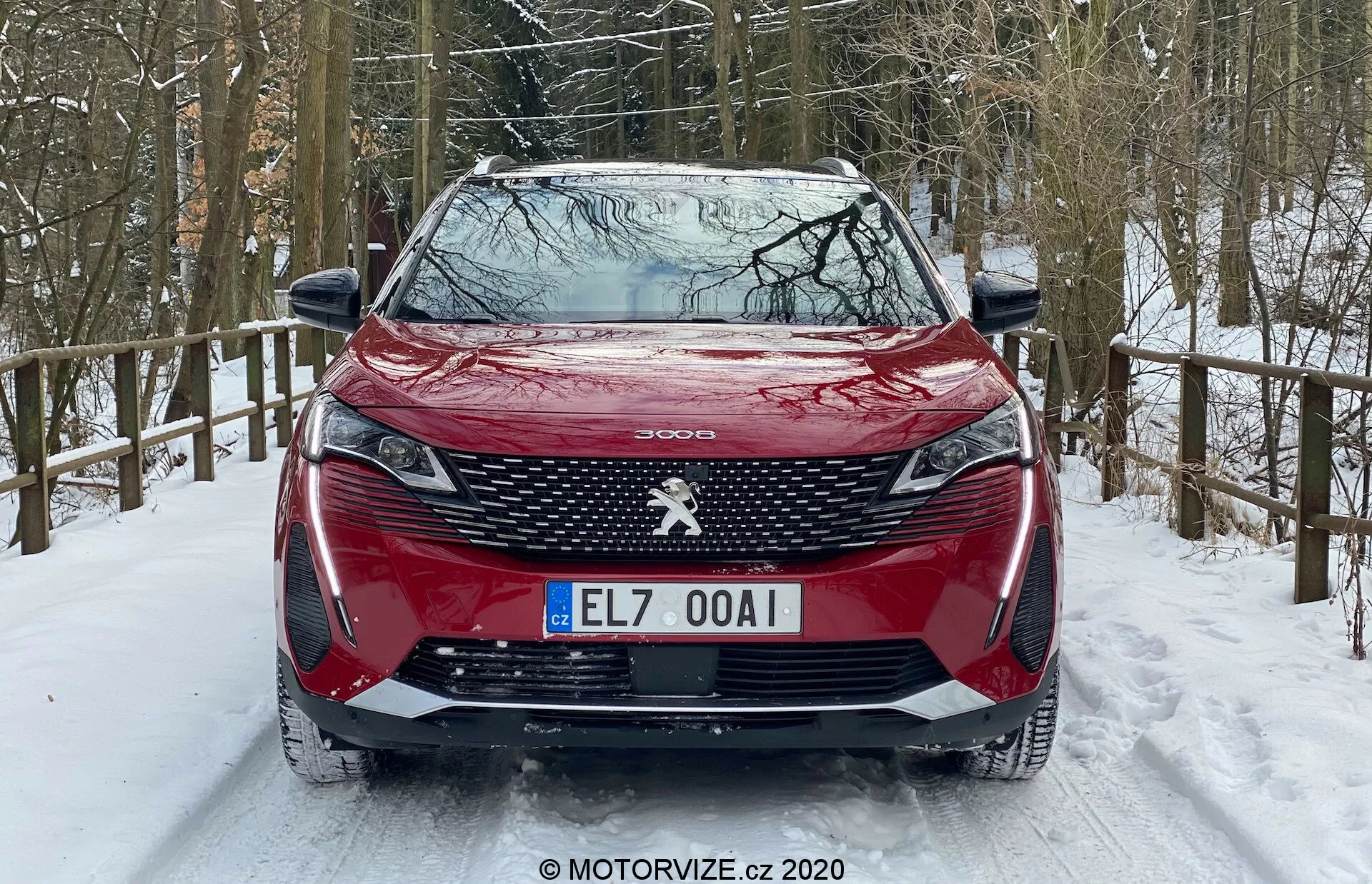 Sprednji pogled na rdeč Peugeot 3008 2021, parkiran na zasneženi cesti, z značilnim vzorcem rešetke, osrednjim emblemom Peugeot, ostrimi žarometi z dnevno svetlobo v obliki zob, oblikovanim pokrovom motorja, sprednjim odbijačem z vgrajenimi parkirnimi senzorji in zasneženo gozdno okolico.