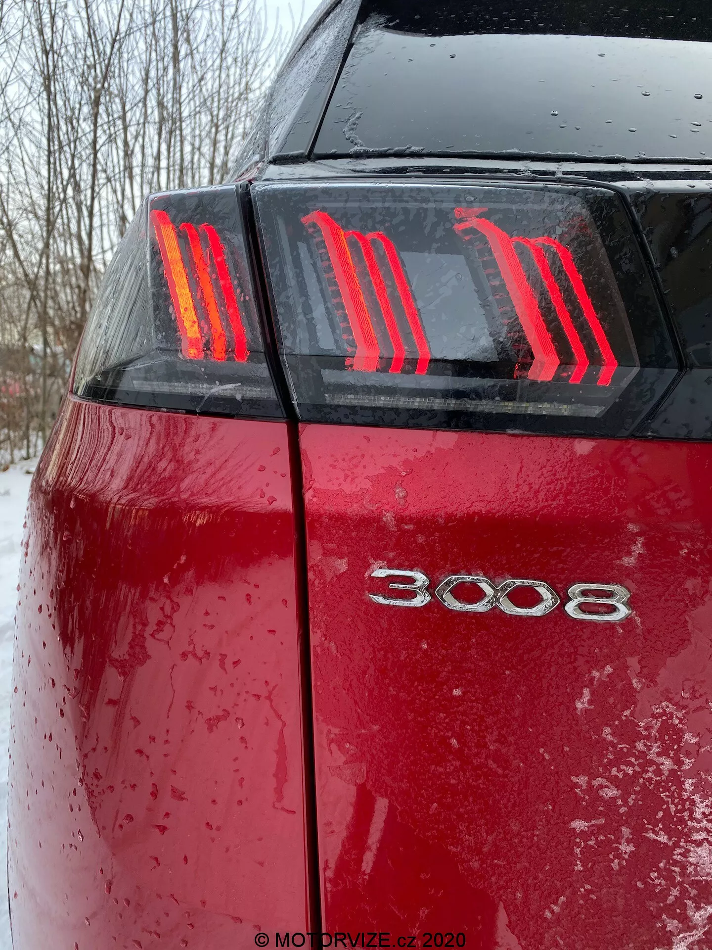 Bližnji posnetek zadnjega dela rdečega Peugeot 3008 2021 iz nizkega kota, ki prikazuje značilne tri navpične LED luči v obliki krempljev in oznako modela '3008'. Mokra površina avtomobila, prekrita z kapljicami vode in umazanijo ali snegom, poudarja nedavno uporabo v slabih vremenskih razmerah pri oblačnih svetlobnih pogojih.