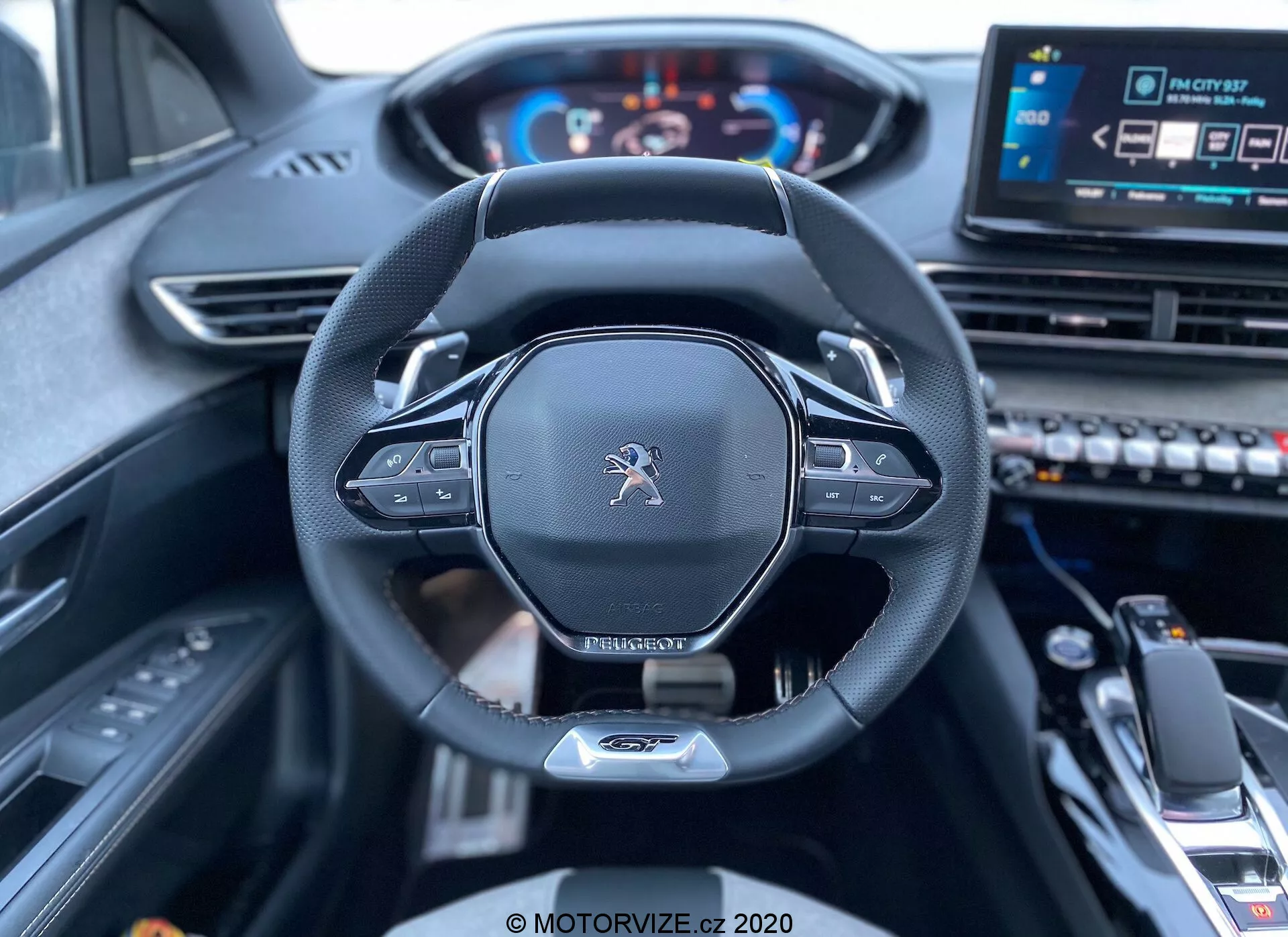 Pogled na notranjost iz voznikovega sedeža Peugeot 3008 2021, ki prikazuje volan in armaturno ploščo. Slika se osredotoča na osrednjem okviru volan, ki ima logotip Peugeot, moderen dizajn s ploščatim spodnjim delom, usnjen rob in nameščene kontrole, verjetno za nastavitve avdia in tempomata. Za volanom so vidne ročice za ročno prestavljanje. Digitalni instrumentni panel prikazuje hitrostomer in tahometer, ki sta osvetljena, kar pomeni, da je vozilo vklopljeno. Na desni strani volana je delno viden osrednji zaslon za informativno-razvedrilni sistem na dotik in različne kontrolne gumbe, skupaj s prestavno ročico in sredinskim naslonom za roke v spodnjem ospredju, kar nakazuje moderen, tehnološko napreden avto z vrhunskim notranjim prostorom.