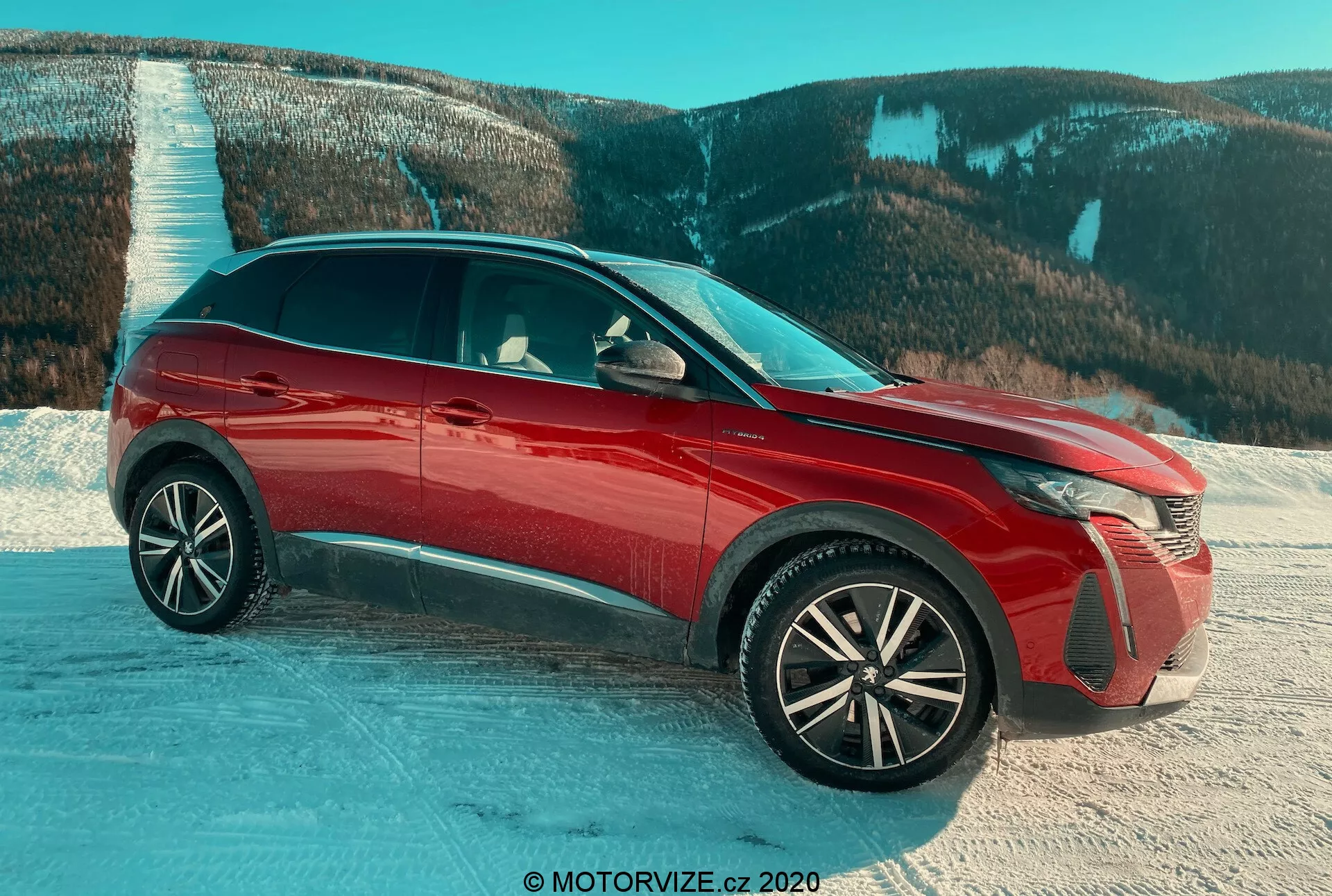 Peugeot 3008 SUV 2021 v rdeči barvi, zajet iz trikotnega sprednjega pogleda na zasneženi površini z gorskim ozadjem, poudarjajoč napredne LED žaromete, zapletene aluminijaste kolesa, sijoč zaključek, aktivne rešetke hladilnika in bočna ogledala z vgrajenimi smerniki, poudarjajoč oblikovalske funkcije in primernost za zahtevne pogoje.