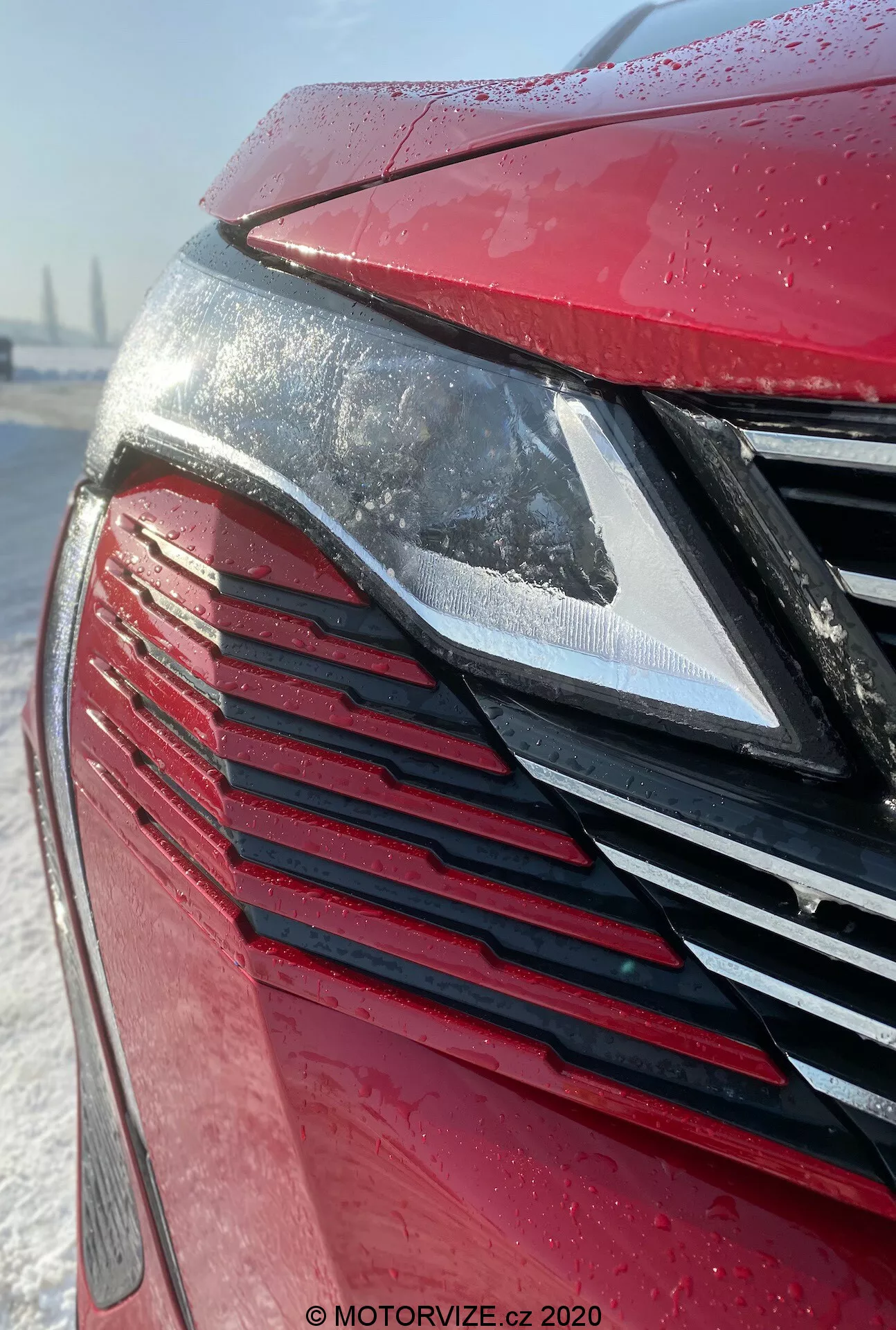Bližnji pogled na sprednji del vozila Peugeot 3008 2021, s poudarkom na sodobni LED-žarometi in maski z navpičnimi lamelami v kovinsko rdeči barvi, prekrit z vodnimi kapljicami, kar namiguje na nedavni dež ali mraz. Snežno ozadje nakazuje na zimske razmere ali detajlno nego vozila.