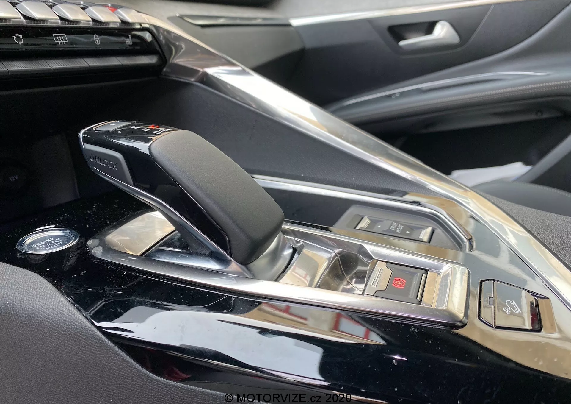 Pogled od blizu na sredinsko konzolo in območje prestavne ročice Peugeot 3008 2021 s strani sopotnika. Predstavlja sodobno, elegantno avtomatsko prestavno ročico z usnjenimi in kromiranimi poudarki, obdano z visokim sijajem konzole. Vidni so gumbi za različne funkcije vozila in vpogled v informacijski sistem, kar nakazuje vrhunsko notranjost avtomobila z visokokakovostnimi materiali in prefinjenimi tehnološkimi funkcijami.