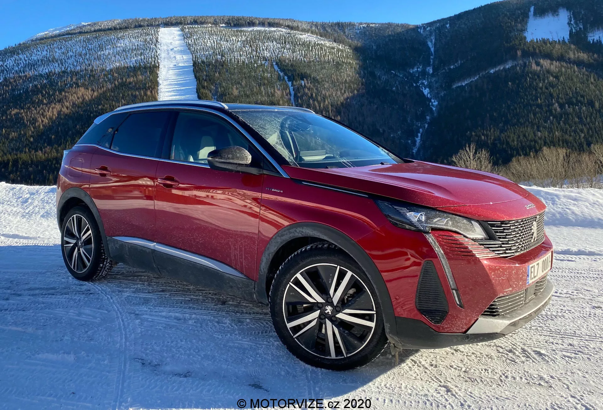 Slika modernega avtomobila, verjetno Peugeot 3008 letnik 2021, posneta iz prednje tričetrtinske perspektive. Kot prikazuje značilno zasnovo LED žarometov avtomobila, mrežo v obliki glavnuka in emblem na sprednji strani. Sprednji odbijač prikazuje sekcije za zajem zraka in ohišja meglenk. Pokrov motorja ima izklesane linije, ki prispevajo k aerodinamiki, medtem ko široko vetrobransko steklo zagotavlja dobro vidljivost naprej. Večkrake aluminijaste platišča nakazujejo bolj športen videz in višjo stopnjo opreme. Elegantna linija strehe in dinamične linije karoserije namigujejo na crossover ali SUV. Vidna so tudi stranska ogledala z vgrajenimi smerniki ter bočne obloge ali okrasni elementi. Prizorišče je zasnežena cesta z gorskim ozadjem, kar nakazuje, da se vozilo lahko spopade z različnimi tereni.