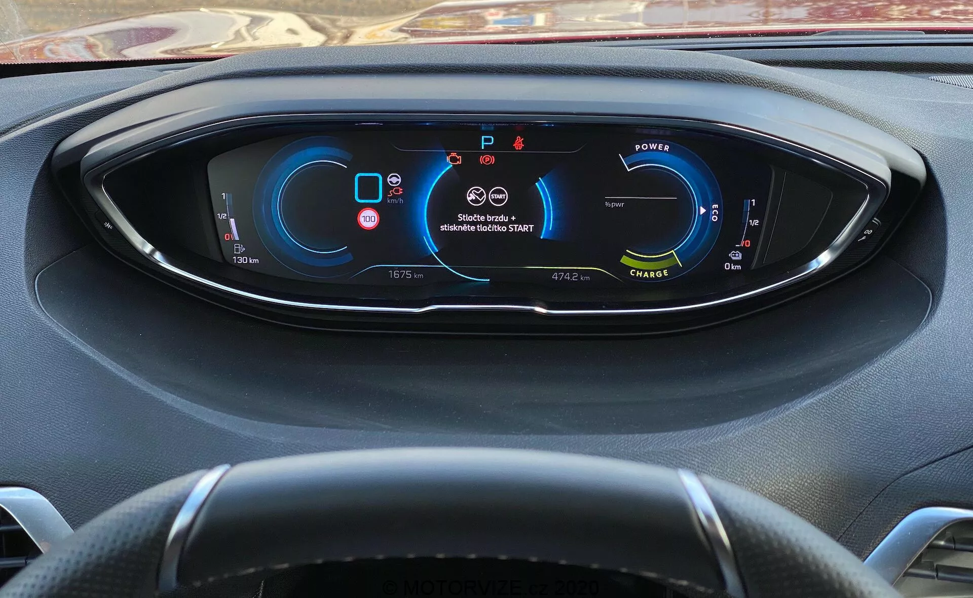 Pogled voznika na instrumentno ploščo Peugeot 3008 iz leta 2021 s popolnoma digitalnim zaslonom, ki prikazuje merilnike in kazalnike vozila. Na levi strani je merilnik moči, ki nakazuje električno/hibridno vozilo; na sredini zgoraj opozorili za varnostni pas in zračne blazine; na desni strani je merilnik hitrosti, ki prikazuje, da je vozilo v mirovanju; spodaj so prikazani menjalnik (parkiranje), števec kilometrov, zunanja temperatura, načini vožnje in razpoložljiv doseg. Delno viden trikraki volan. Pogled izza volana z rahlim nagibom navzdol.
