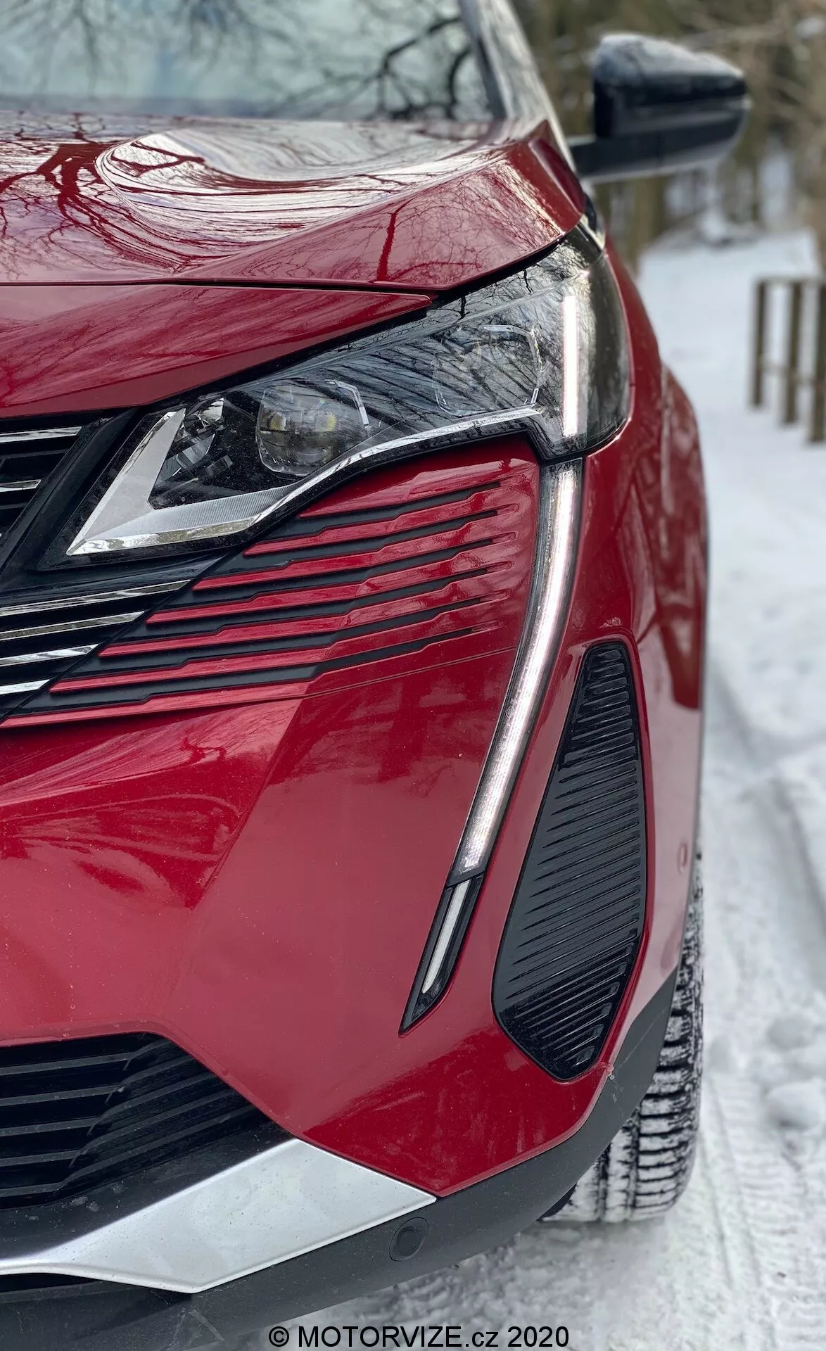 Od blizu z nizkega kota ujeta podoba modernega rdečega Peugeot 3008 iz leta 2021. Poudarek je na sprednjem levem delu, ki prikazuje elegantne kotne LED žaromete, veliko agresivno rešetko, izrazit dizajn ventilacijskih odprtin, sijajni lak in ostro oblikovane karoserijske plošče. Stransko ogledalo ima integrirane smerne kazalce, scena pa vključuje zasneženo področje, kar nakazuje zimske razmere. Viden vodni žig MOTORVIZE.cz.
