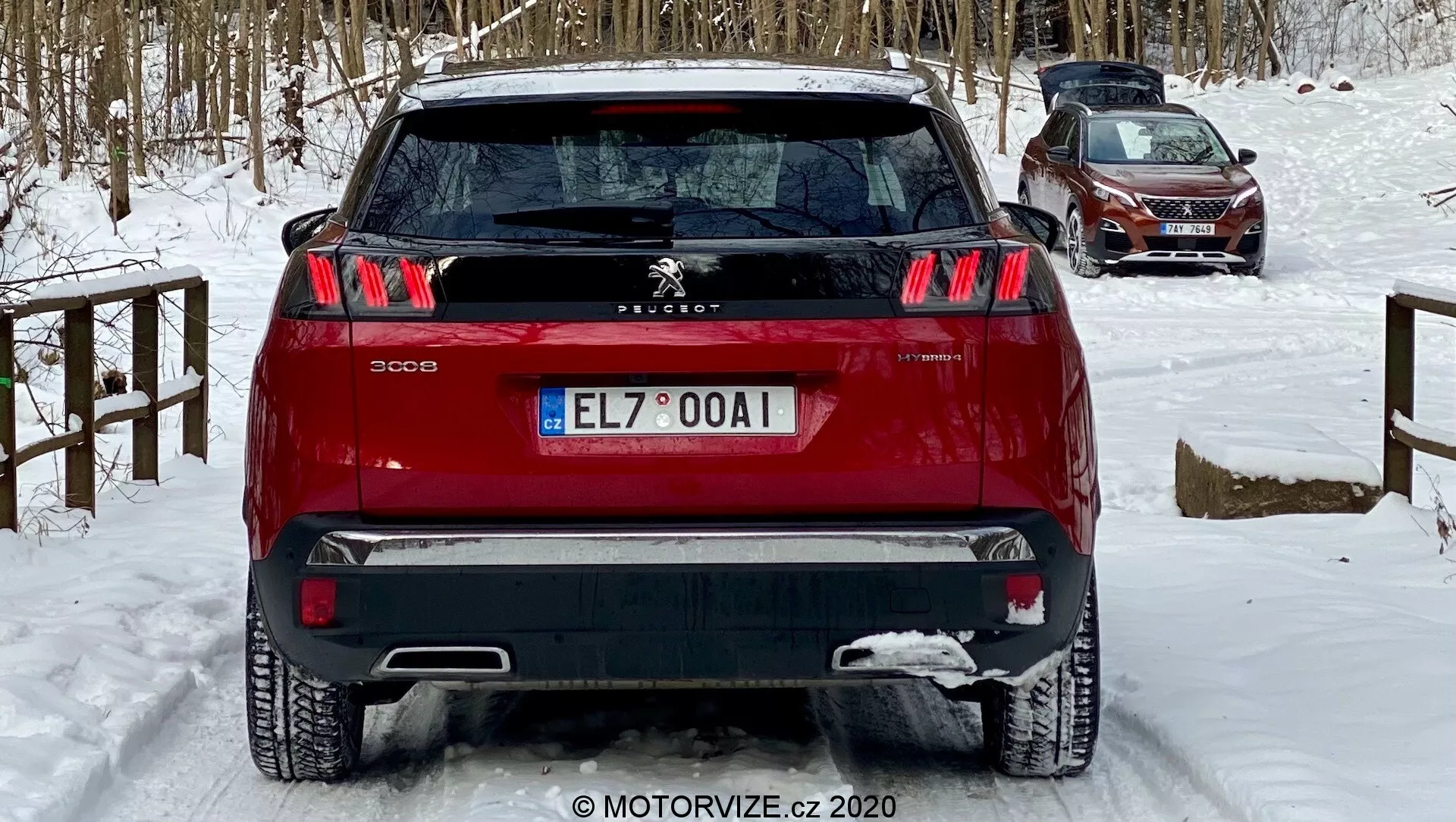 Zadnji pogled na rdečega Peugeot 3008 SUV posnet iz rahlega nizkega kota, ki poudarja njegove značilne zadnje luči s tremi črtami, osrednji emblem Peugeot Lion in dvobarvni odbijač. Vozilo, nameščeno na zasneženem terenu, nakazuje zimske pogoje. Vidne lastnosti vključujejo zadnje parkirne senzorje, zadnji brisalec vetrobrana in zgornjo zadnjo anteno, kar nakazuje na tehnologijo za pomoč vozniku in potencialno funkcionalnost GPS/radio, primerno za razprave o avtomobilskem oblikovanju, varnostnih funkcijah in zimskih vožnjah.