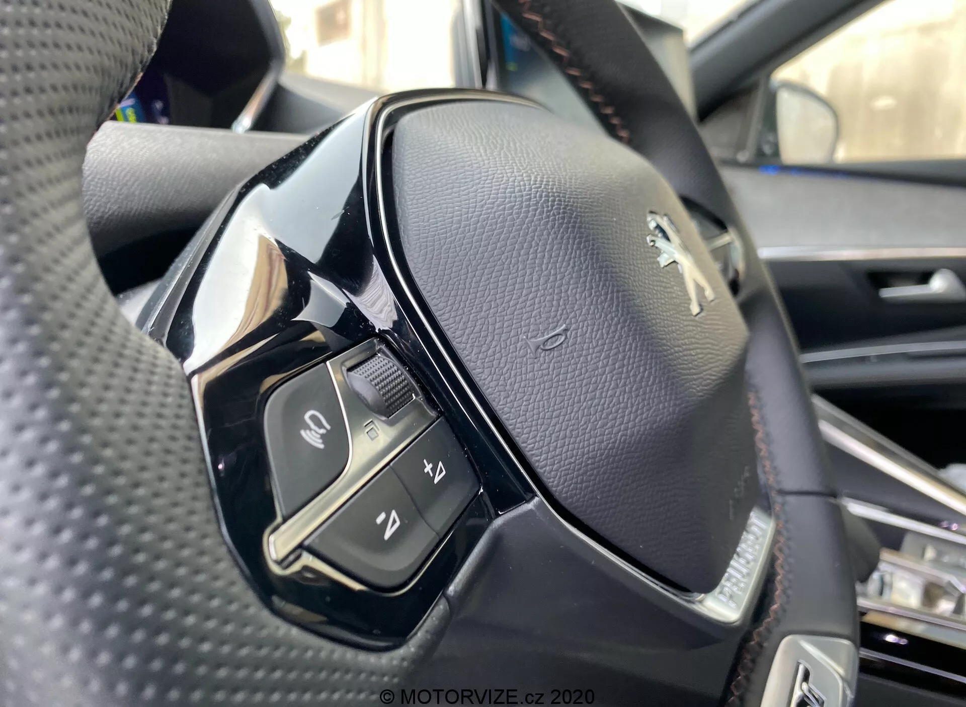 Bližnji pogled na volanski obroč iz perspektive sedeža voznika v Peugeot 3008 2021. Slika poudarja levo stran volana, posneta iz rahlo nizkega kota in prikazuje podrobnosti levi del krmila in Peugeot logotip. Volanski obroč je prevlečen s črnim perforiranim usnjem, z mešanico črnih mat in sijajnih materialov. Vidni kontrolni elementi vključujejo stikalo, gumbe za funkcije medijev in komunikacije, kot je sprejem ali zaključek klicev, aktivacijo glasovnega upravljanja in še eno neidentificirano funkcijo. Ostrina je visok, prikazujejo šivke na usnju in sijajni zaključek na nekaterih plastičnih delih, morda tipke na dotik.