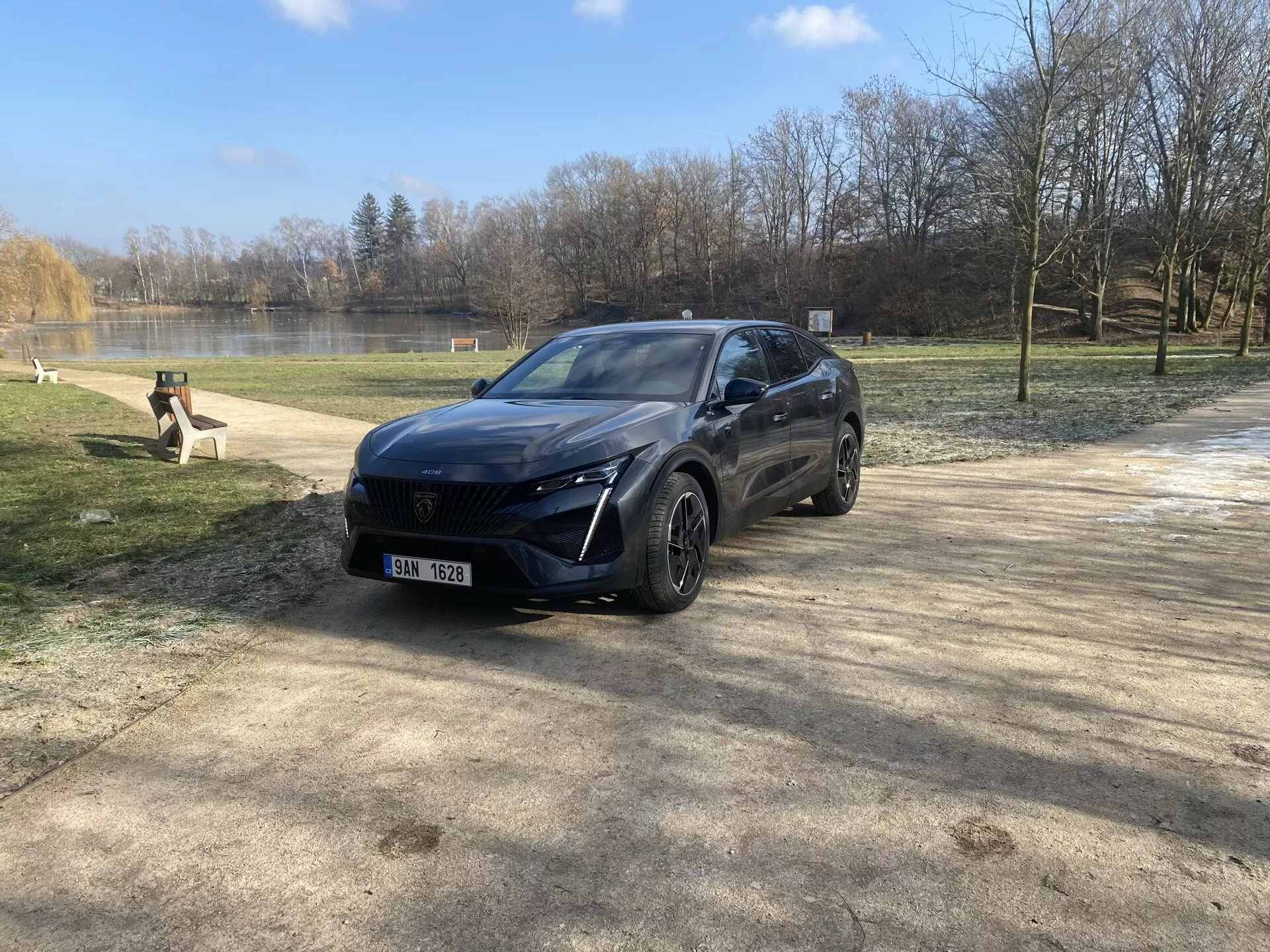 Peugeot 408 letnika 2023 parkiran v naravnem okolju pozimi ali zgodaj spomladi, predstavljajoč njegov sodoben crossover dizajn, LED žaromete, sistem sprednjih senzorjev, zlitinska kolesa in premijski zaključek iz pogleda sprednjega tričetrtinskega kota pod sončno svetlobo z bleščanjem leče.