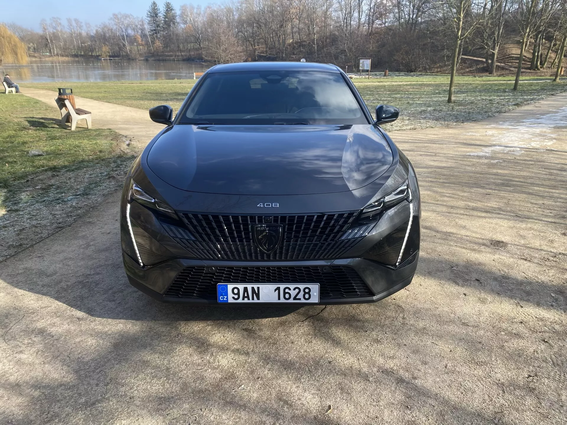 Sprednji pogled na Peugeot 408 letnik 2023, parkiran na prodnati površini z ozadjem dreves in vode, poudarja njegovo veliko rešetko z diamantnimi vzorci, elegantne LED žaromete, izklesane linije pokrova in napredne funkcije pomoči vozniku