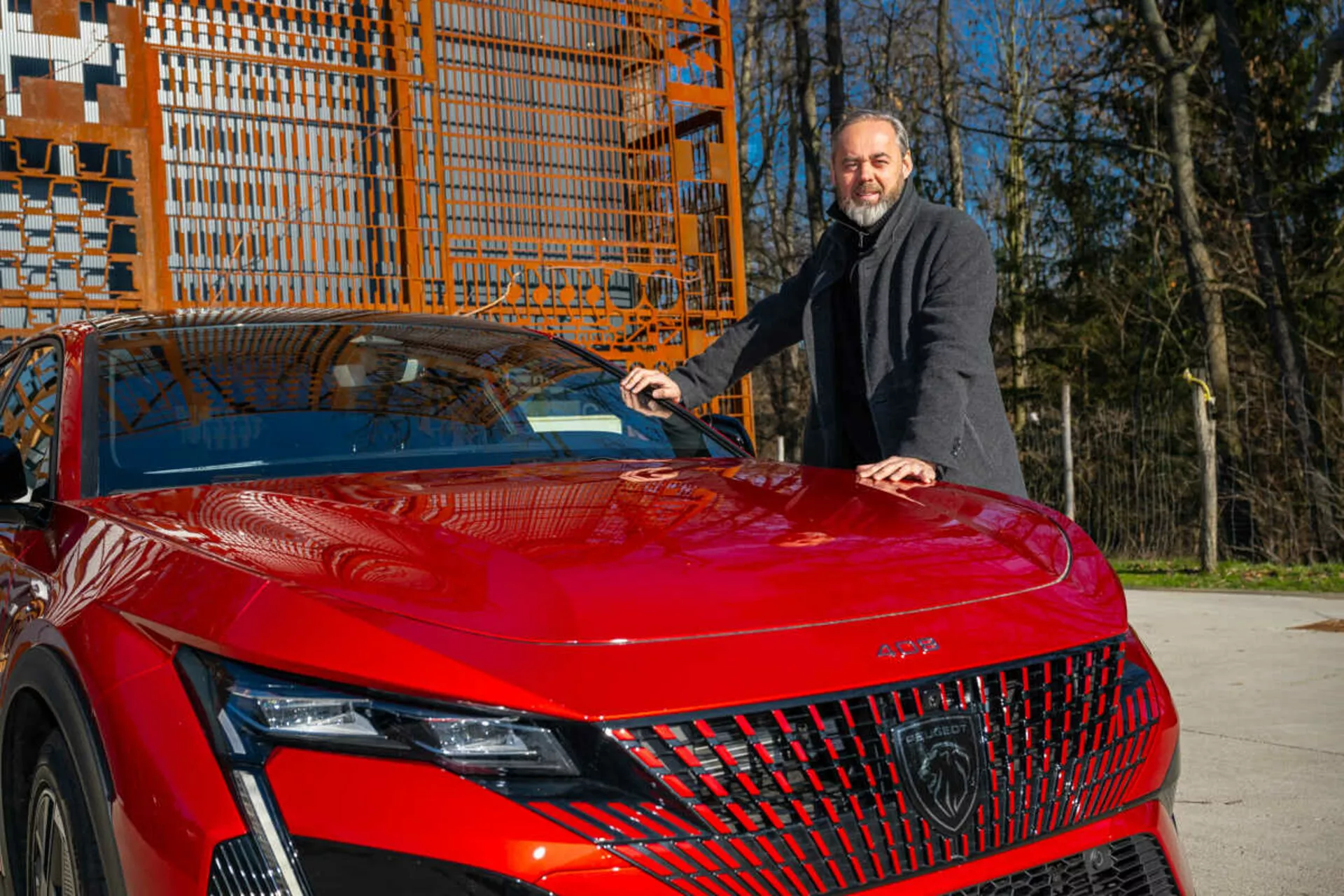 Rdeč model Peugeot 408 leta 2023 prikazuje svojo sprednjo levo četrtino na dnevni svetlobi, poudarjajoč podrobne LED žaromete, geometrično rešetko in športno, aerodinamično pokrov motorja z moškim v temnem plašču ob strani, ki roko položi na pokrov