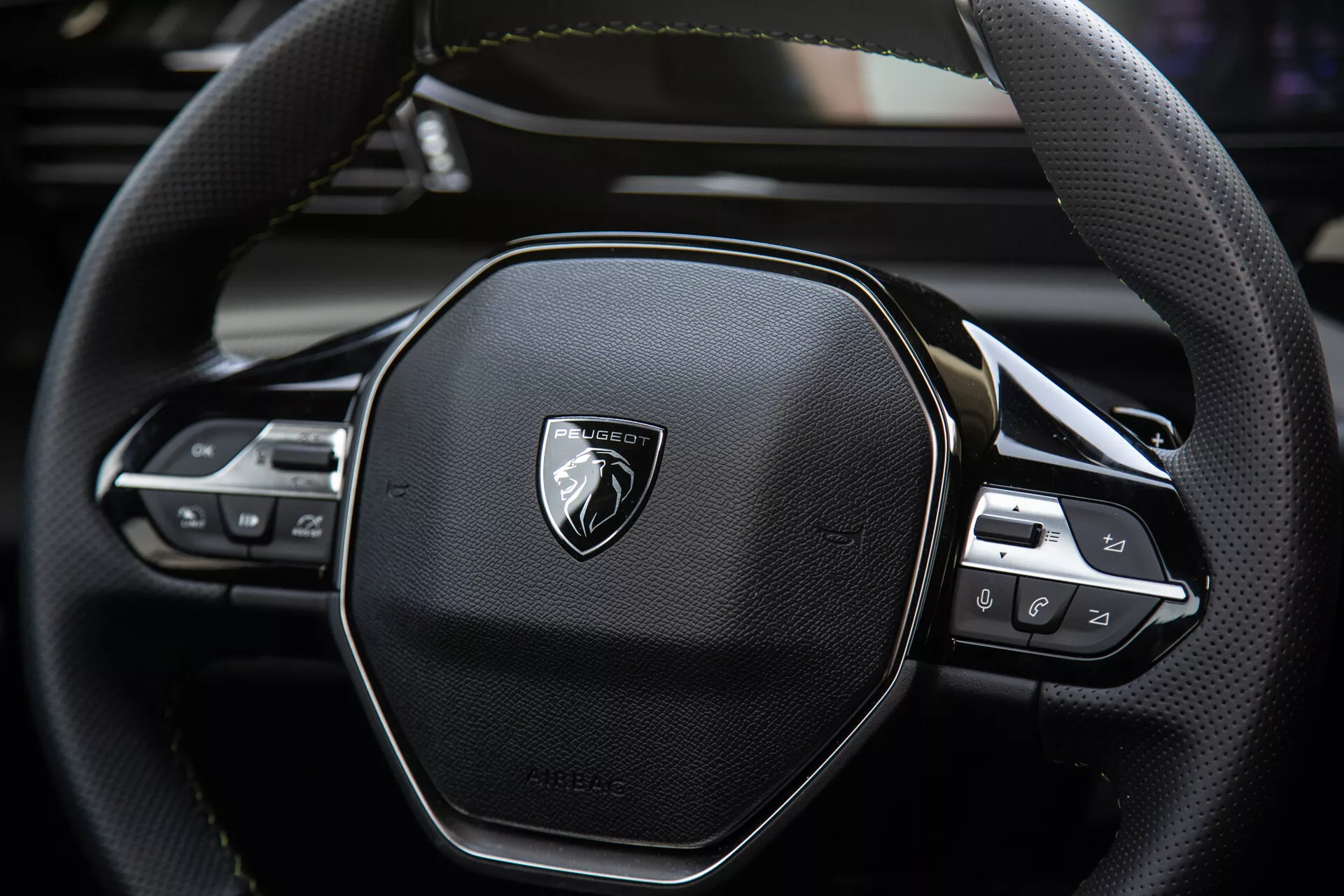 Podroben pogled na volan Peugeota 408 2023 ki prikazuje ergonomično šestkotno oblikovanje, šivanje iz usnja, vgrajene kontrolnike tehnologije za avdio in nastavitve tempomata, z emblemo na brušenem kovinu.