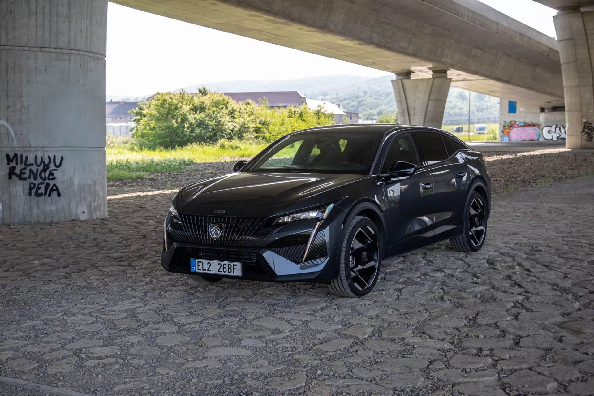 Peugeot 408 2023, posnet z zornega kota tri četrtine, ki poudarja njegove elegantne LED žaromete, vzorčasto masko in športni aerodinamični sprednji odbijač pod naravno dnevno svetlobo na tlakovani površini pod nadvozom