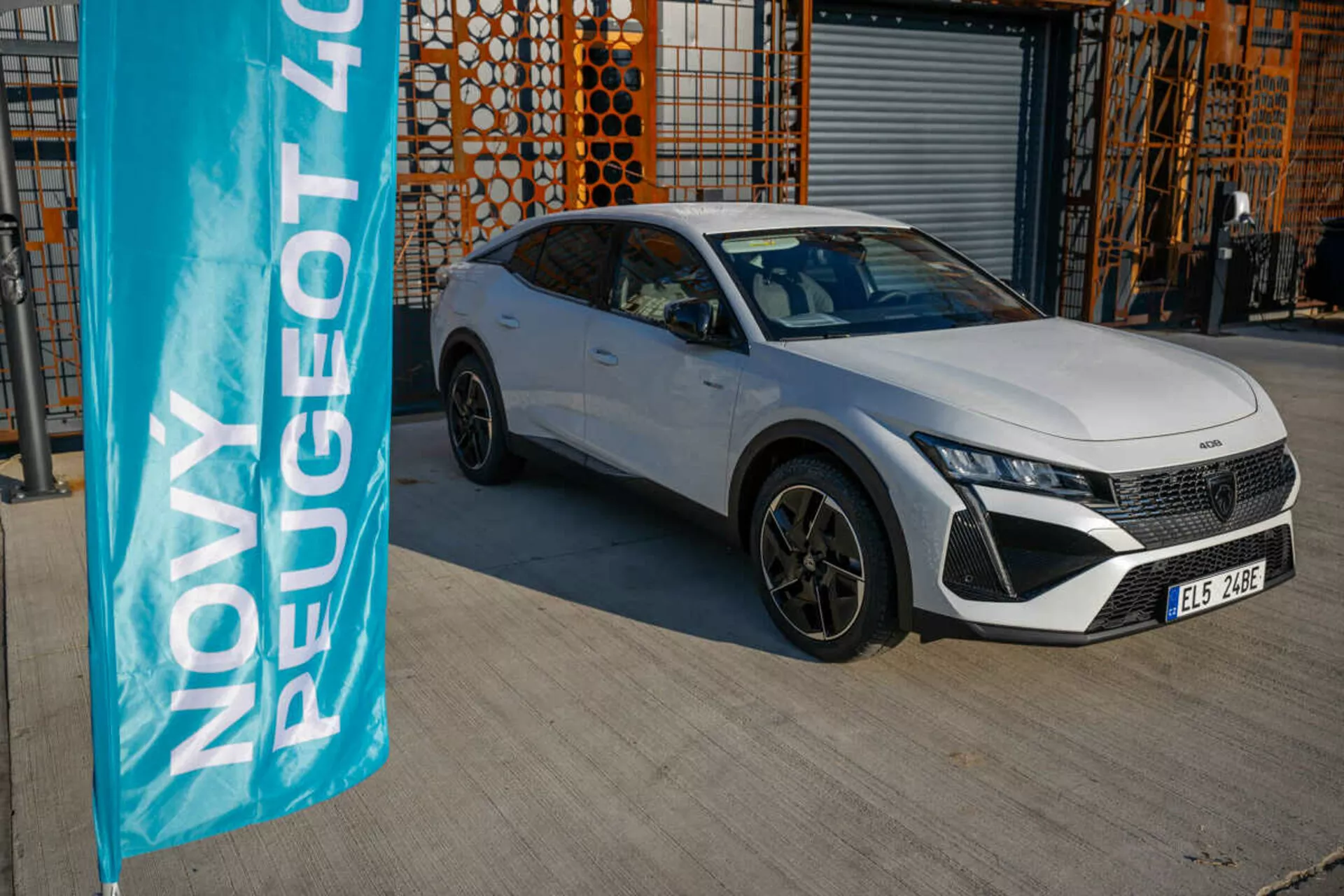 Beli Peugeot 408 letnik 2023 parkiran na prostem z industrijsko arhitekturo v ozadju, ki predstavlja oblikovalske in tehnološke značilnosti vozila pod naravno dnevno svetlobo