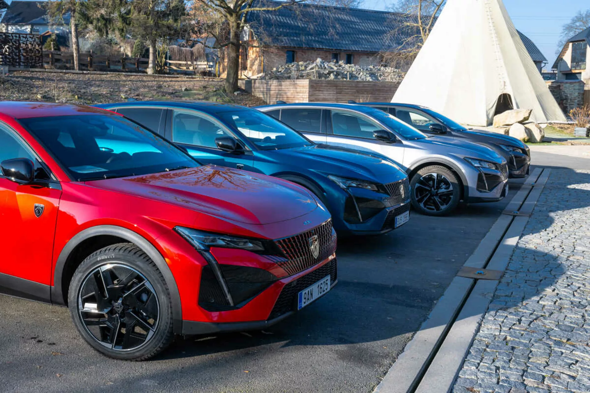 Linija vozil Peugeot 408 letnik 2023 z rdečim avtomobilom v ospredju s športnimi večkrakimi kolesi in LED žarometi, ki nakazujejo na električne ali hibridne modele, parkirana na tlakovani ob kamniti površini