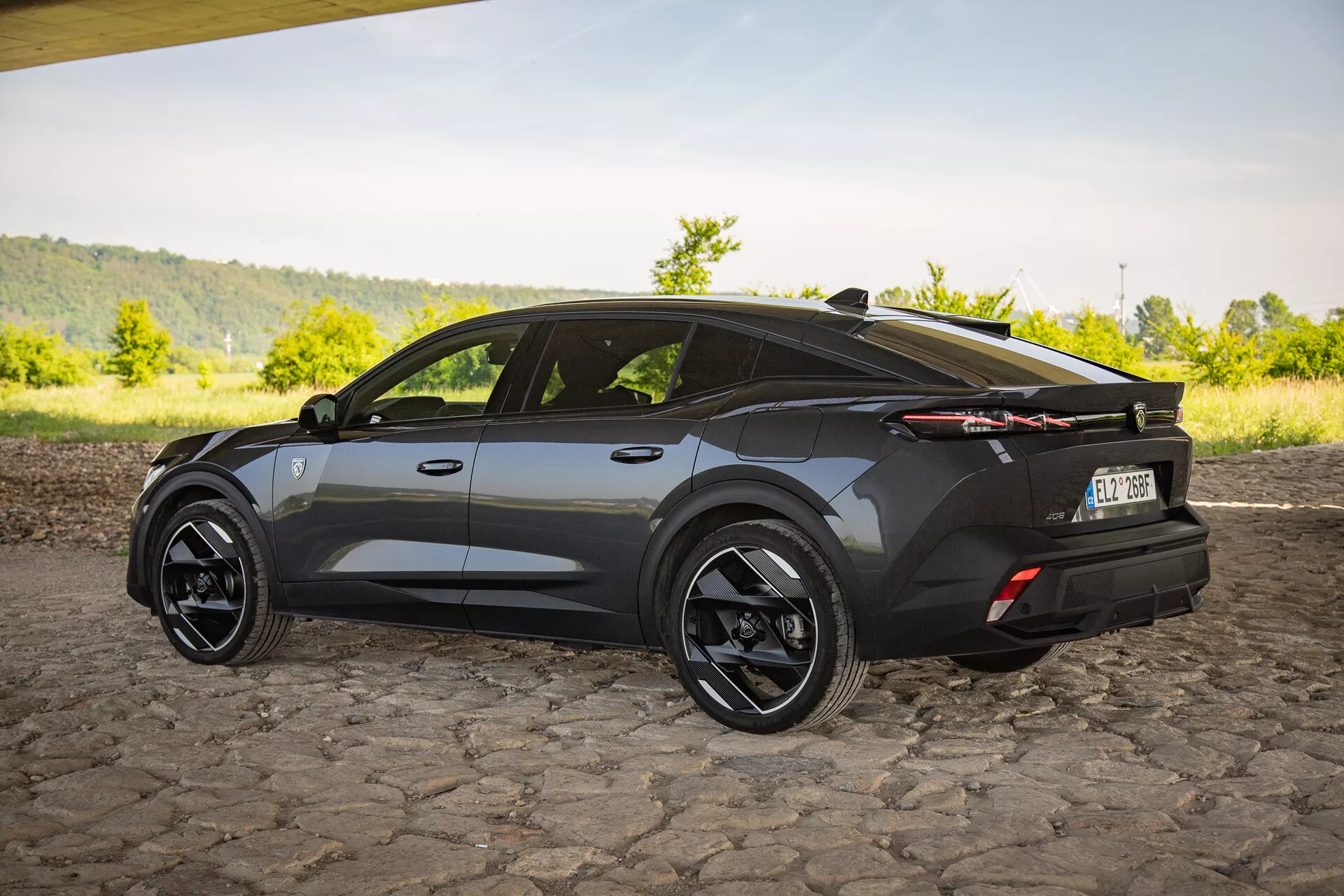 Crossover SUV Peugeot 408 letnika 2023 v temnem zaključku, ki z zornega kota tri četrtine od zadaj poudarja aerodinamično oblikovanje z ostrimi koti, športno poševno streho, velika črna lita kolesa, značilne LED zadnje luči in diskreten zadnji spojler na tlakovani površini