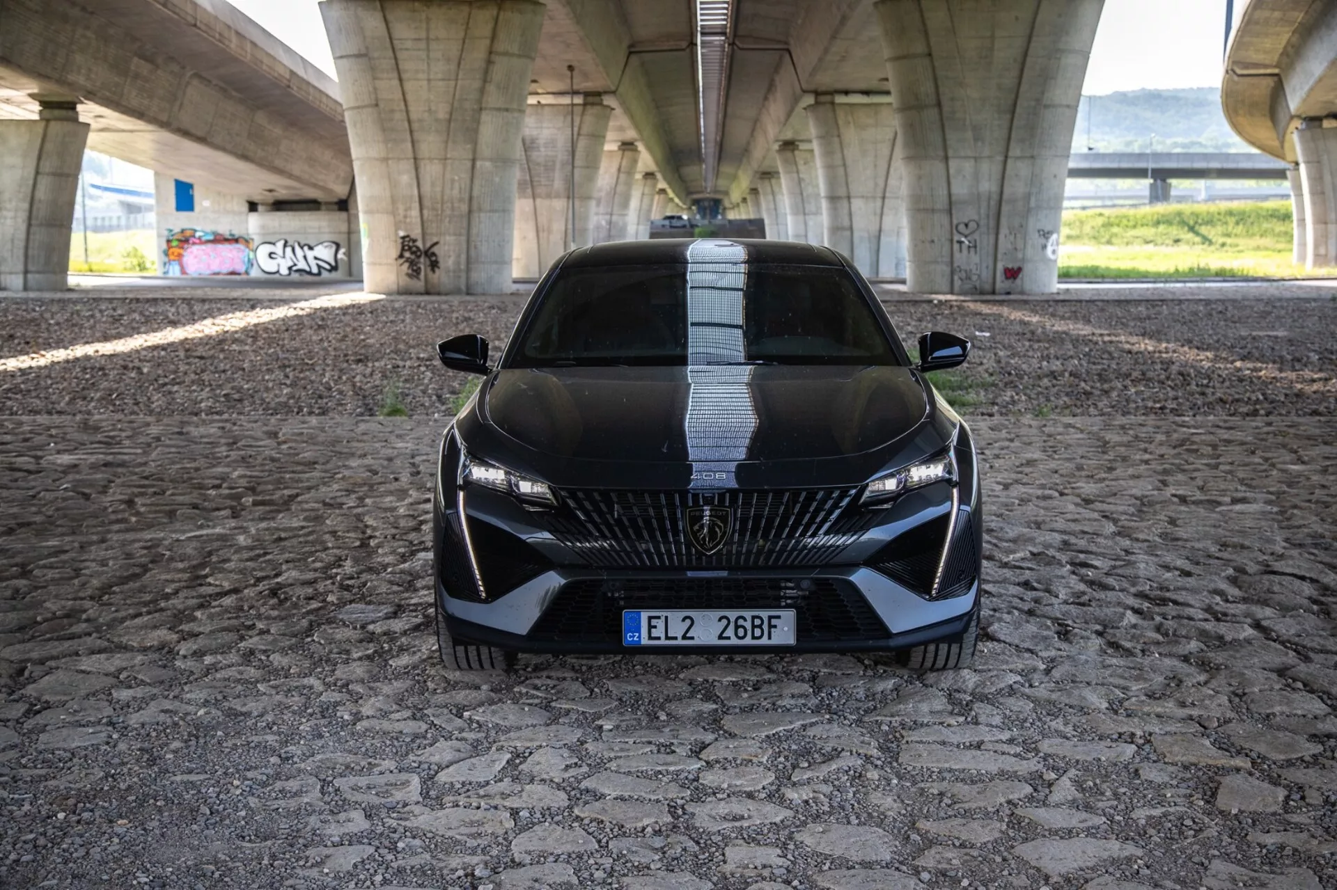 Peugeot 408 iz leta 2023 v dinamičnem pogledu od spredaj, ki poudarja njegovo agresivno držo in športno oblikovanje z detajlno rešetko, elegantnimi LED žarometi in aerodinamičnimi značilnostmi na vzorčasti tlakovci pod mestnim mostom