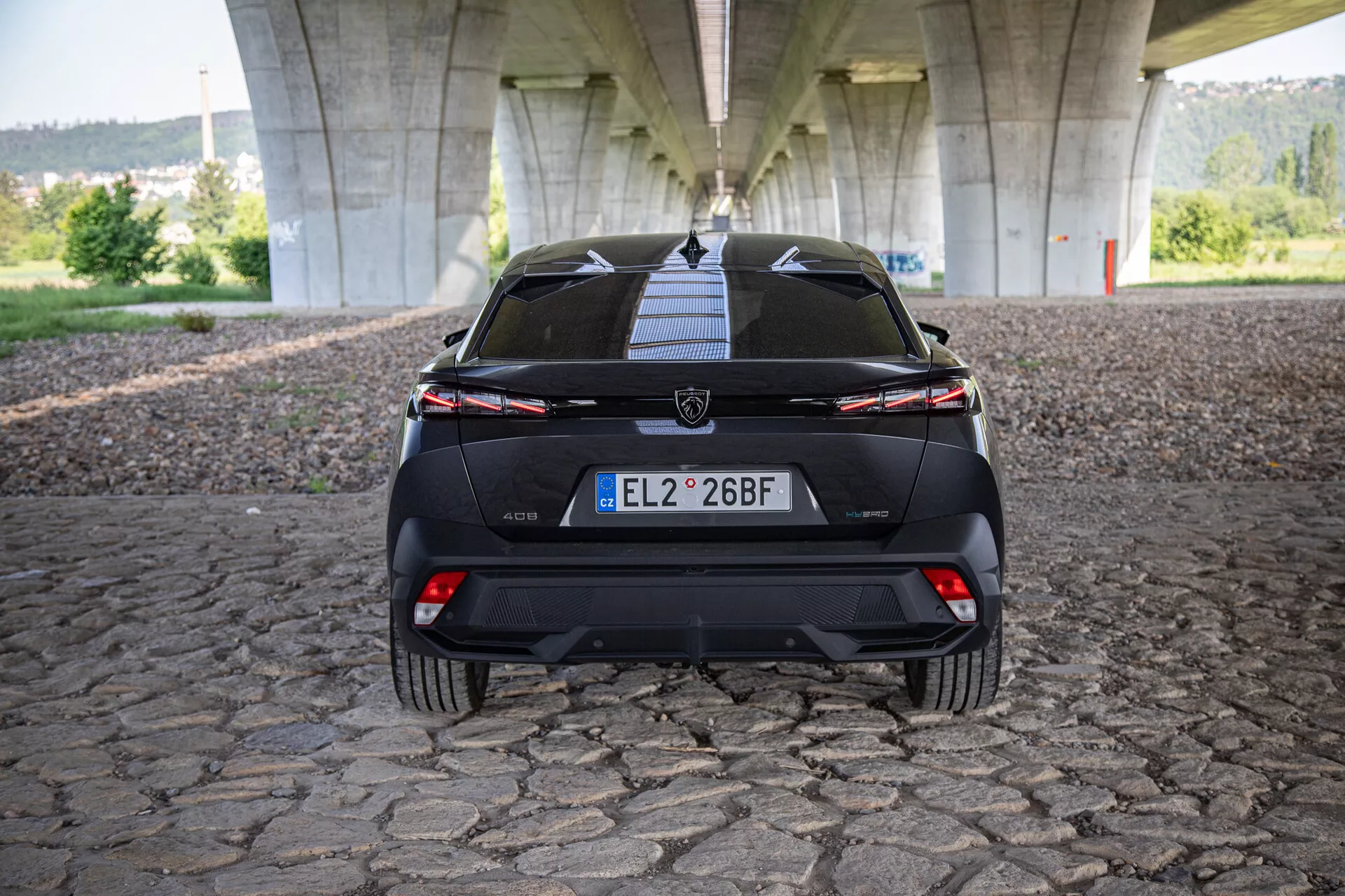 Zadnji pogled na Peugeot 408 2023, ki prikazuje prtljažna vrata, odbijač, spodnji difuzor, zadnje luči s povezovalno svetlobno letevjo in senzorje za pomoč pri parkiranju, fotografirano pod mostom na tlakovani površini