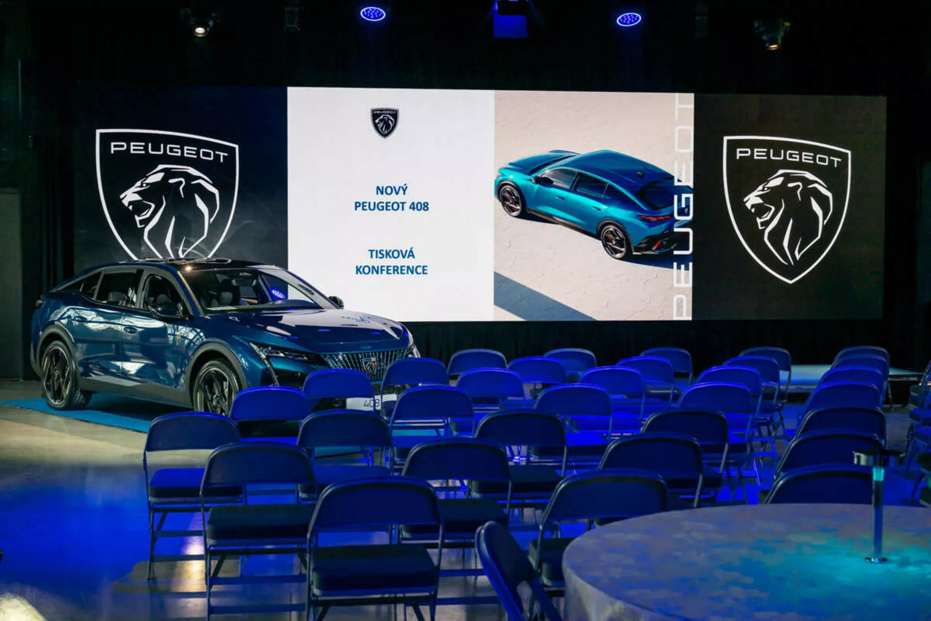 Peugeot 408 prikazan na tiskovni konferenci, pogled pod 45-stopinjskim kotom poudarja njegov sprednji in stranski profil, elegantno obliko in LED žaromete, z digitalnimi zasloni in blagovno znamko v ozadju