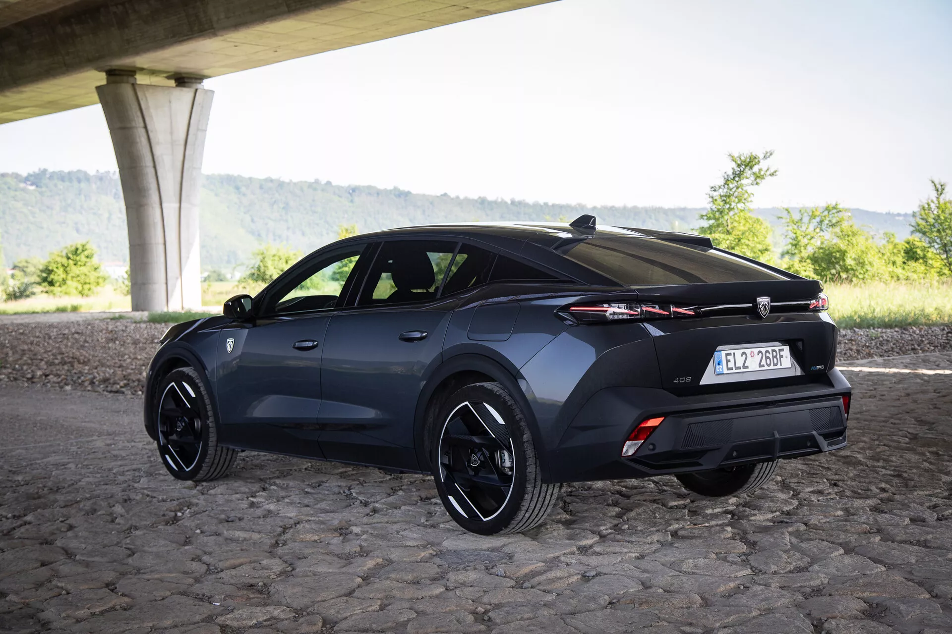 Tričetrtinski zadnji pogled na hatchback Peugeot 408 letnik 2023 pod mostom na tlakovcih s športnimi platišči in napredno svetlobno tehnologijo