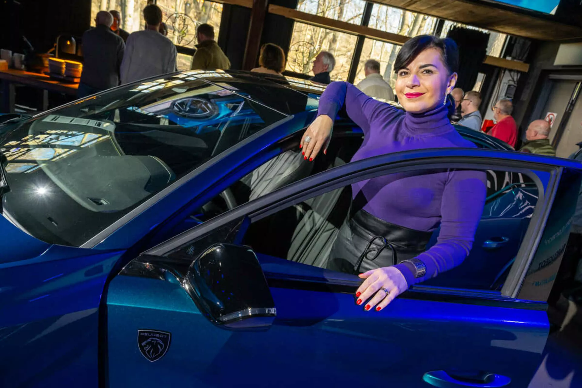Živahen modri športni avtomobil, verjetno Lamborghini, posnet iz kota treh četrtin, ki poudarja njegove elegantne linije, ostre LED žaromete in ikonični logotip v obliki ščita, z osebo, ki naključno interagira z vozilom, kar nakazuje na luksuz in slog.